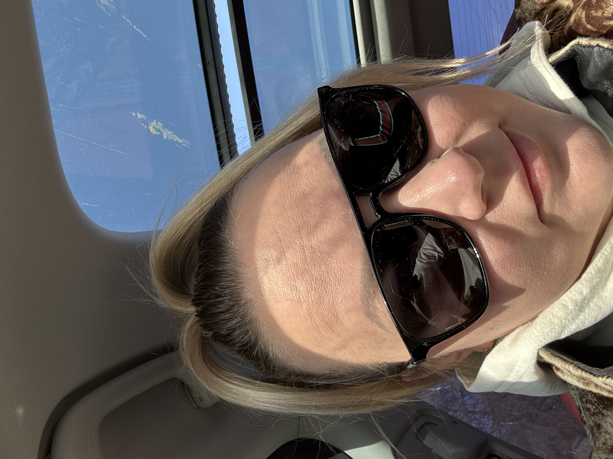 The image depicts a close-up portrait of an adult female taken inside a vehicle. She has blonde hair pulled back into a ponytail and wears black-framed sunglasses, which reflect a distinct red and green striped pattern on the right lens. She is dressed in a light-colored high-collared garment beneath a brown-toned patterned jacket. Strong sunlight illuminates her face from the upper left, casting defined light and shadow. The vehicle's interior headliner is visible at the top left of the frame. A sunroof above the subject reveals a clear blue sky with discernible streaks or smudges on the glass. Bare tree branches are visible through a side window on the viewer's right. The subject's mouth exhibits a slight upward curve, indicating a gentle smile. This scene is identified as taking place in the city of Хохряки, Russia, though no specific visual markers within the image confirm this precise geographical location.
