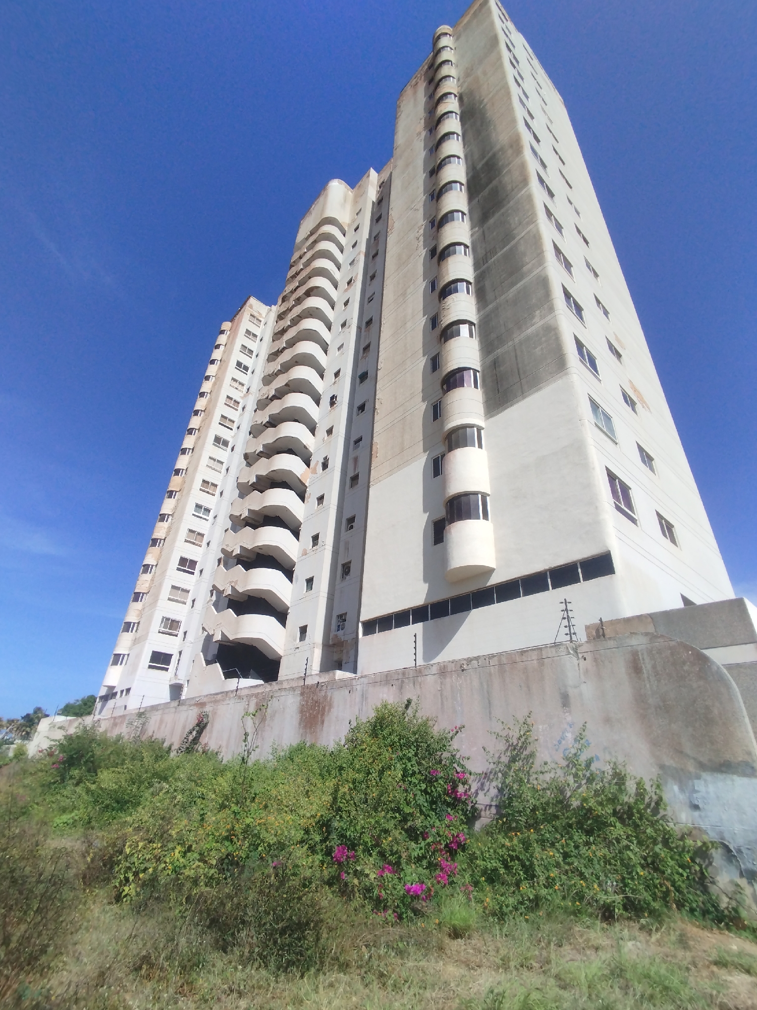 The image depicts a tall, weathered concrete building set against a bright blue sky. The building is located in Porlamar, Venezuela. The structure is multi-story, with a noticeable vertical emphasis. It features a series of balconies on one side, and a more simple, rectangular facade with windows on the other. The exterior shows signs of age and weathering. The building is set on a raised foundation, with vegetation growing at its base. A concrete wall and some fencing are visible between the vegetation and the building itself.