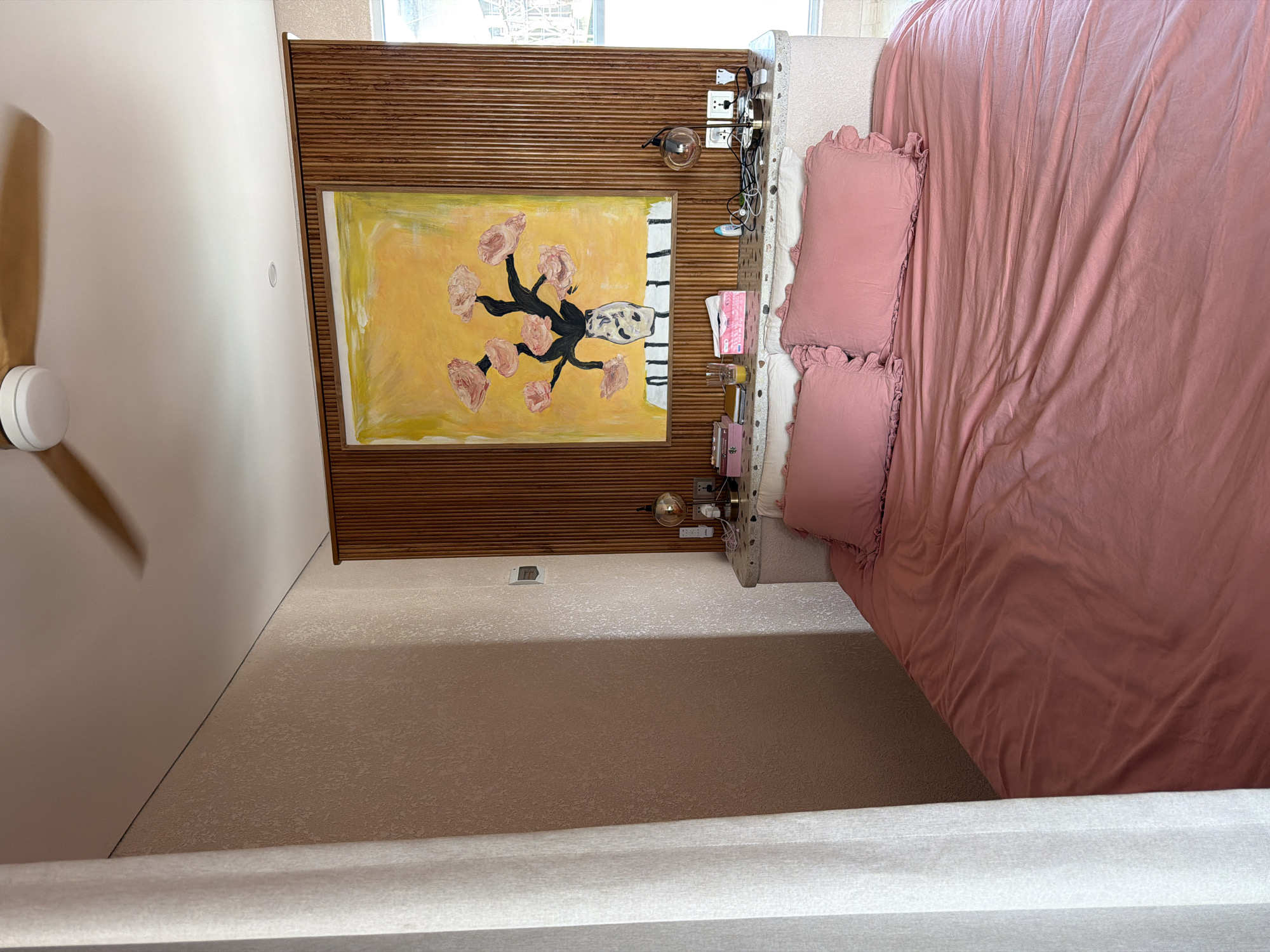 A top-down perspective captures a bedroom scene. A bed with pink bedding and pillows dominates the right side of the frame. Adjacent to the bed is a headboard constructed of horizontal wooden slats with a painting hanging in the center. A bedside table is attached to the headboard, holding lamps, books, and various items. The room's walls are painted light pink, with a ceiling fan visible in the upper left corner. The floor appears to be carpeted in a neutral color. The location is possibly in Mengwi, Indonesia.