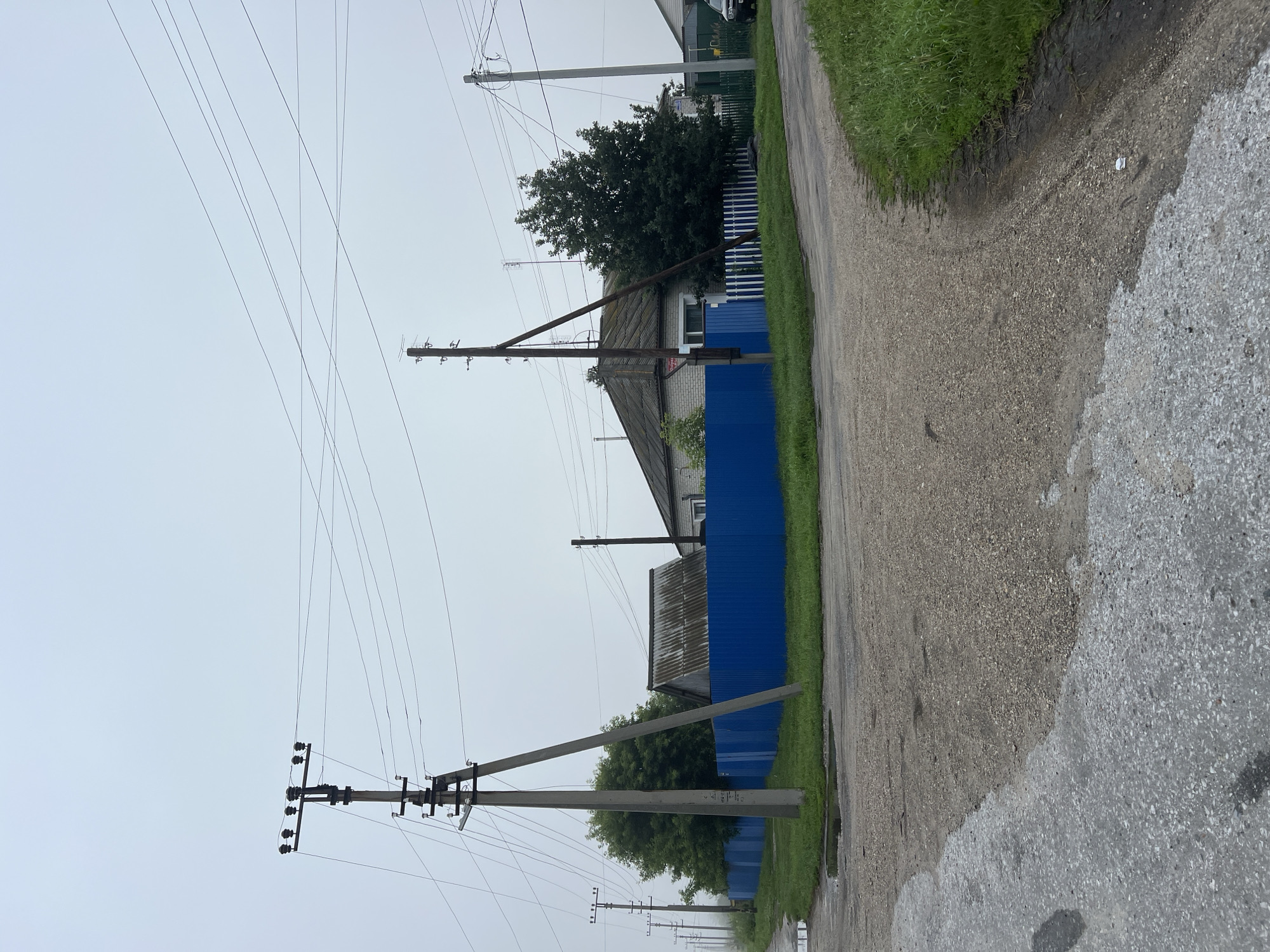 This is a medium-shot, eye-level, vertical photograph of a rural intersection under a hazy, overcast sky in Pivkino, Russia. A gravel and asphalt road, wet with puddles, forks in the foreground. To the left, a tall, concrete power pole with multiple insulators and crossbars stands at the edge of a grassy verge. Beyond the verge, a bright blue metal fence runs horizontally. Behind the fence, a low-slung building with a corrugated metal roof is visible, partially obscured by a large, leafy green tree. Another, darker power pole stands further down the road. To the right of the intersection, the road curves away, bordered by lush green grass. More power lines crisscross the sky. The overall atmosphere is muted and somewhat damp.