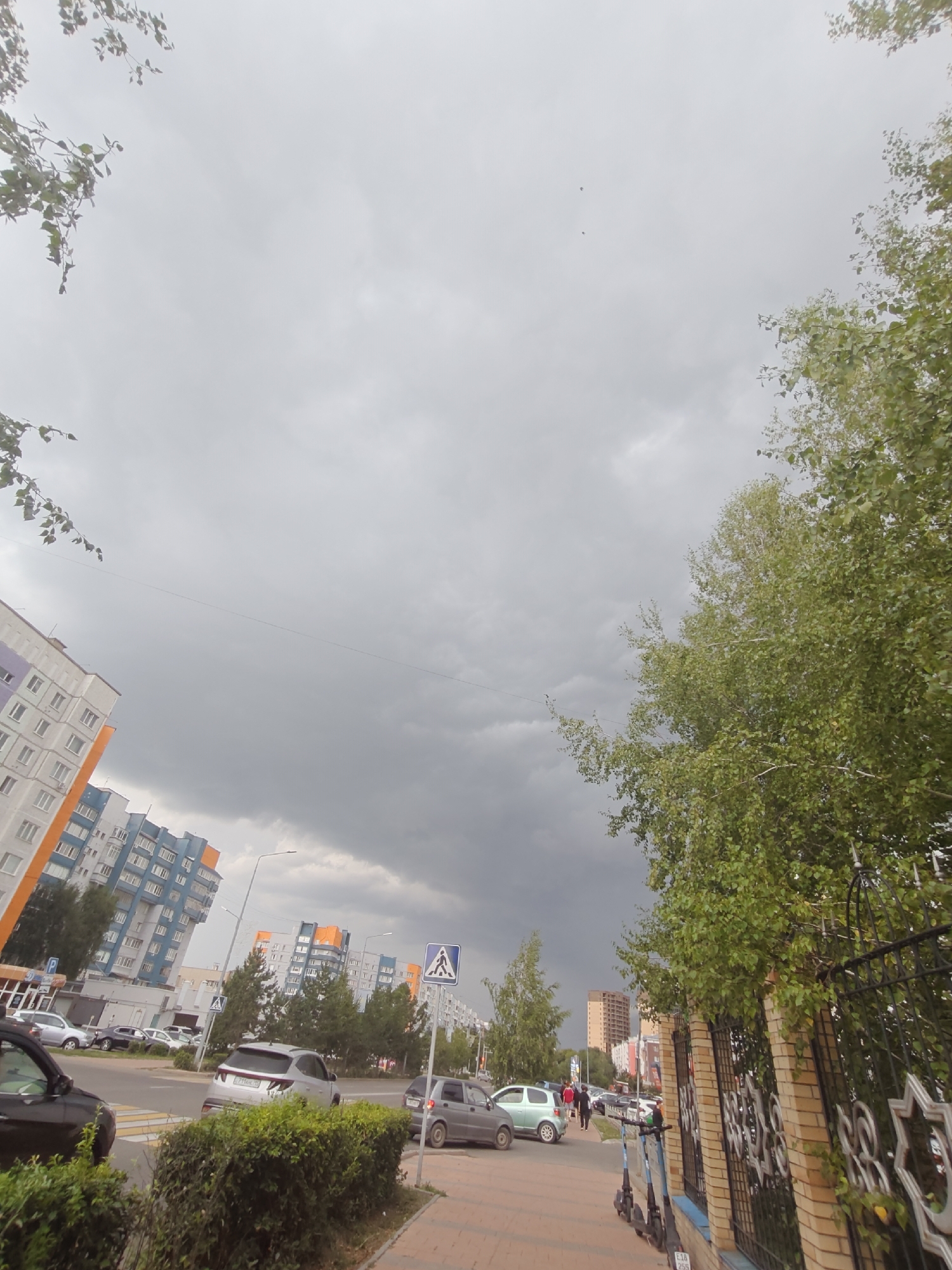 The image captures a street scene in Petropavl, Kazakhstan, under an overcast sky. On the left, a tall, multi-story apartment building with orange and white facades is visible, characteristic of urban residential architecture. Adjacent to it are more buildings of similar design, featuring blue and orange accents. A street with parked cars runs horizontally across the mid-ground, with a crosswalk marked by yellow and white stripes. Several vehicles, including a silver SUV, a light-colored sedan, and a dark car, are present. Pedestrians are seen walking on the sidewalk on the right side of the street. A decorative metal fence with brick pillars runs along the sidewalk, adorned with star-shaped metalwork. Lush green trees with dense foliage frame the right side of the image, partially obscuring the sky. The overall atmosphere suggests an urban environment on a cloudy day.