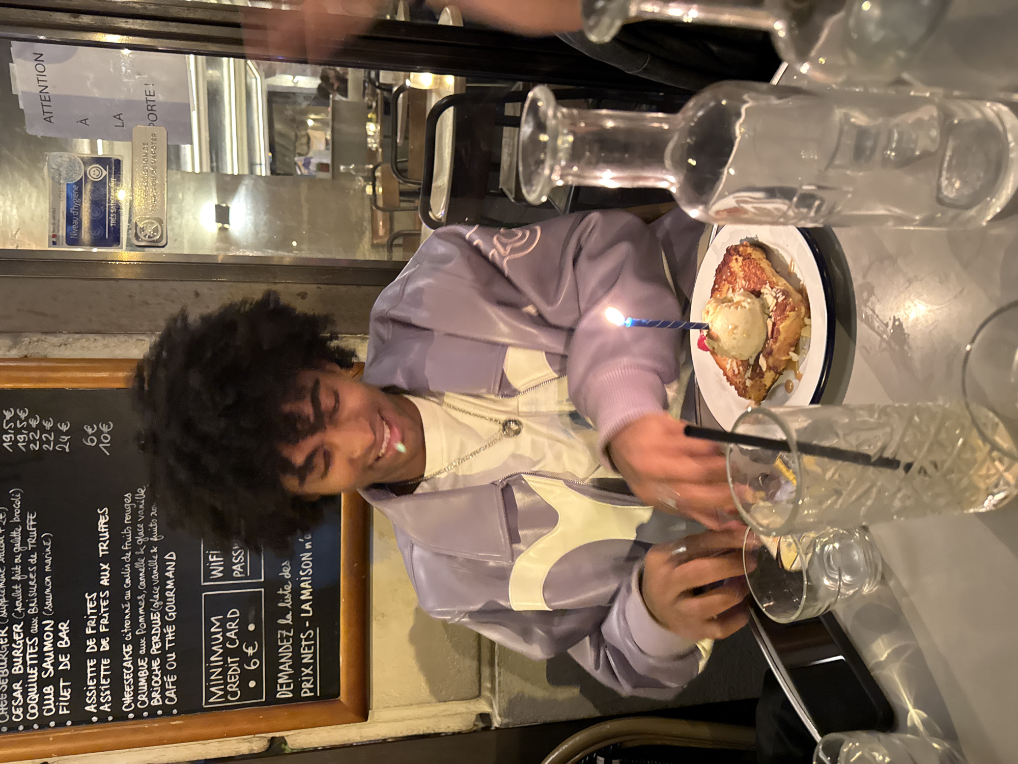 The image depicts a person with curly hair, wearing a purple jacket with white accents and a silver necklace, smiling and holding a black straw over a glass. A plate with what appears to be brioche perdue, topped with a scoop of vanilla ice cream and a lit birthday candle, is visible. Several empty glasses are on the table. A menu board is mounted on the wall showing prices in Euros and contains the following text: "ATTENTION A LA PORTE! Niveau d'hygiène," "CHEESEBURGER (supplement raclette +2€), CESAR BURGER (poulet frit ou galette brocoli), COQUILLETTES aux BRISURES de TRUFFE, CLUB SAUMON (saumon marine), FILET DE BAR, ASSIETTE DE FRITES, ASSIETTE DE FRITES AUX TRUFFES, CHEESECAKE citronné au coulis de fruits rouges, CRUMBLE aux Pommes, cannelle & glace vanille, BRIOCHE PERDUE (glace vanille & fruits ro), CAFE OU THE GOURMAND, MINIMUM CREDIT CARD • 6€, WiFi PASS, DEMANDEZ la liste des PRIX NETS - LA MAISON n'a" and "19,5€, 19,5€, 22€, 22€, 24€, 6€, 10€". Based on the menu's language and the presence of the hygiene rating on the door, the location is a restaurant in Paris, France.