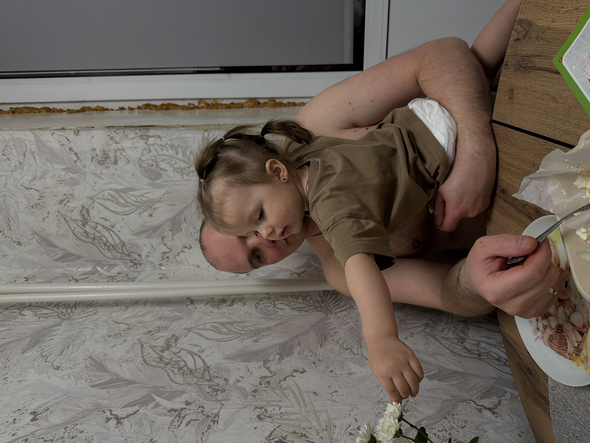 The image depicts a man holding a young child near a table with a plate of food, next to a wall adorned with patterned wallpaper and a window. The man, fair-skinned with short hair, is holding the child with his arms around their torso. The child, dressed in a brown shirt and wearing a diaper, is reaching towards white flowers with their right hand. The man is holding a fork in his right hand, near a plate containing food that includes what appears to be sliced eggs. The background includes a window, the wall, and part of a table.
While there is no definitive confirmation of the location or city using solely the image, it is not possible to determine whether the image was taken in Reshetnikovo, Russia, or any other specific location based on the visual details alone.