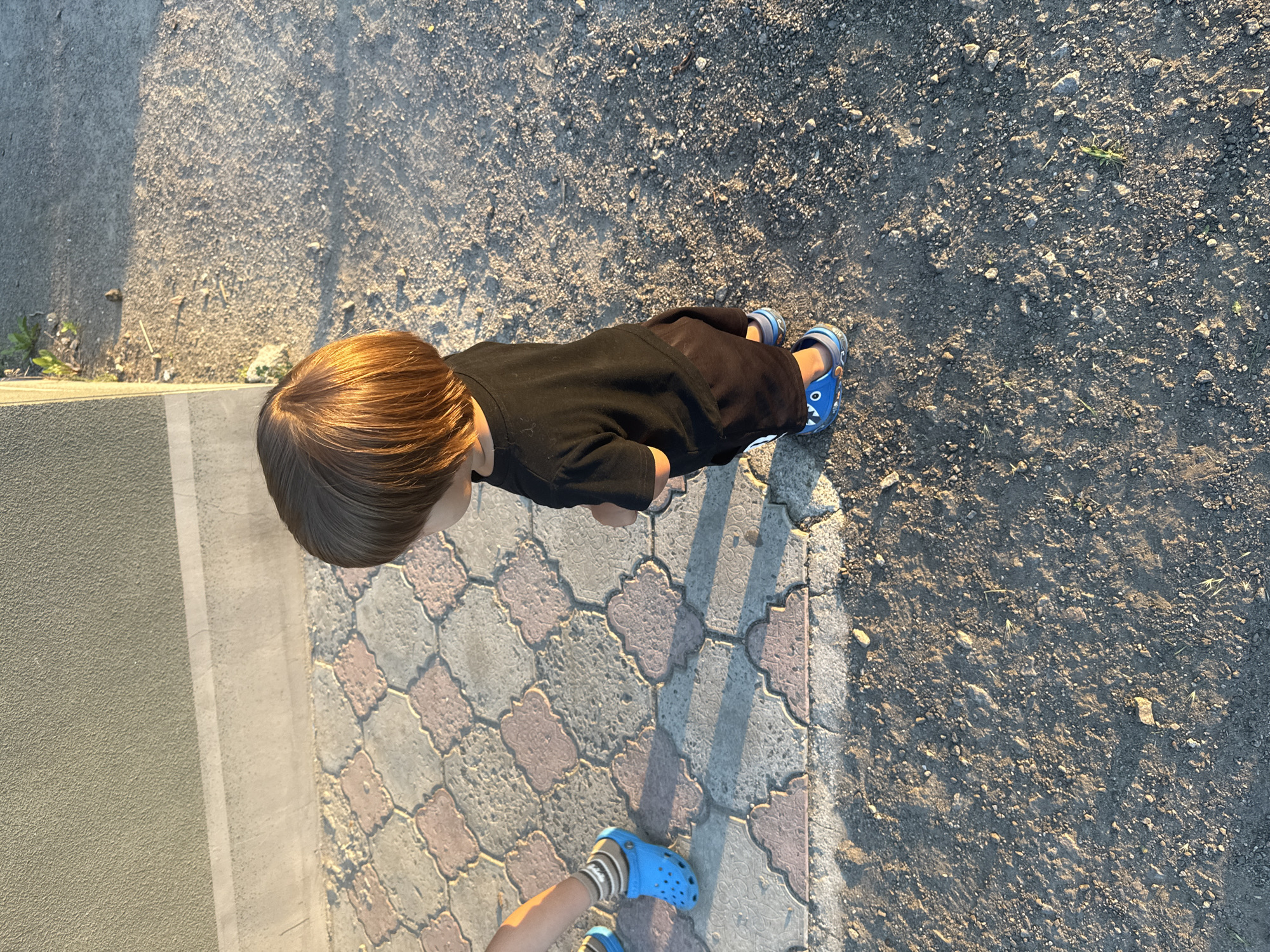 A young child stands on a gravel surface, facing away from the camera and towards a patterned walkway. The child is wearing a black t-shirt, dark shorts, and blue clogs with a cartoonish design. To the left of the child, another child's legs are visible, also wearing blue clogs and striped socks. The walkway is made of interlocking paving stones in shades of beige and pink. Behind the walkway is a textured, light-colored wall. The scene is outdoors, illuminated by the warm light of what appears to be late afternoon or early evening sun, casting shadows. The environment suggests a residential area or a public space within the city of Dnipro, Ukraine, given the time of day and typical urban or suburban landscaping.