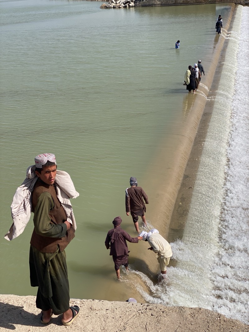 The image captures a scene near Kim, Afghanistan. In the foreground, a young man stands on a concrete ledge, his attire consisting of dark green pants and shirt, a brown vest, sandals, and a patterned head covering. He has a light-colored cloth draped over his left shoulder. To his right, three individuals are wading in shallow water as they traverse a concrete dam. One is wearing a brown outfit and turban, reaching out to assist another person in a light-colored outfit and turban, who appears to be navigating the flowing water. A third person in a brown outfit and turban follows behind. Further along the dam, a group of people dressed in dark clothing is walking along the structure. In the distance, a solitary figure is standing in the water, and another group is near the opposite bank. The water is a murky green, and the dam has water flowing over its edge. The sky is light, indicating daylight.
