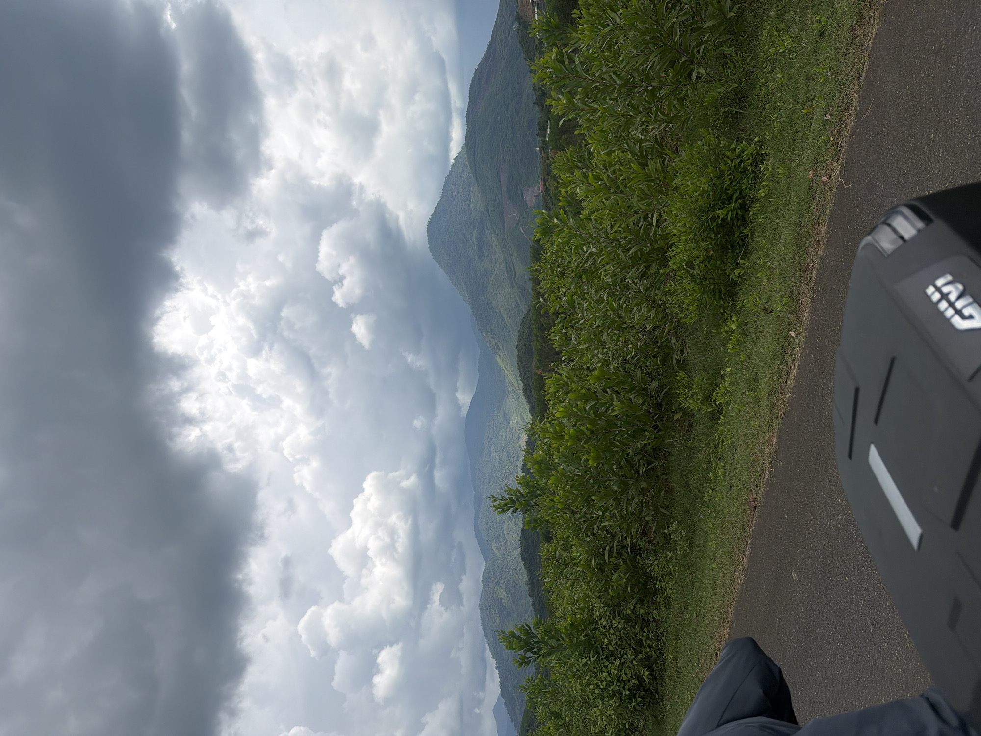 An overcast sky with dramatic clouds hangs over a landscape of lush green hills and mountains. In the foreground, a dark asphalt road runs alongside a dense line of green bushes and vegetation. The bottom of the image shows a portion of a black motorcycle top case, possibly with a Givi logo visible. The scene appears to be taken from a moving vehicle, suggested by the angle and the edge of a person's dark blue clothing on the left. The overall impression is one of a scenic, natural environment in Khanh Vinh, Vietnam, under brooding weather conditions.
