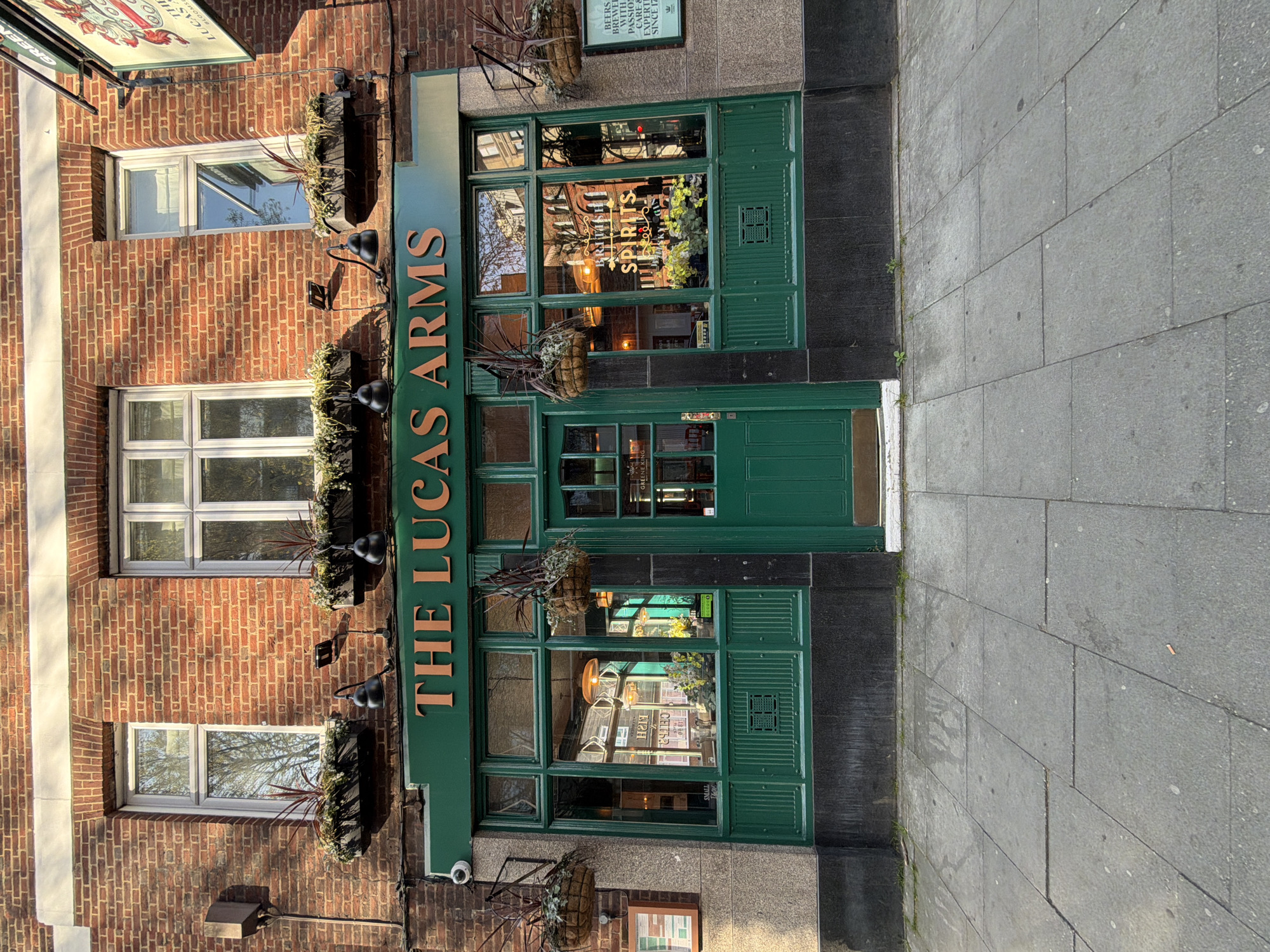 The image shows the exterior of The Lucas Arms, a pub located in London, United Kingdom. The pub is built of red brick and features green trim around the windows and entrance. There are flower boxes with plants under the windows on the first two stories. A green awning above the entrance displays the pub's name in gold lettering. The front windows show reflections and interior details, with signs advertising "British Spirits" and "Greene King" brewery. A sign next to the entrance advertises “Beers Brewer with Passion Care & Expertise since 17”. The image is taken from a ground-level perspective, showing a portion of the sidewalk in the foreground.