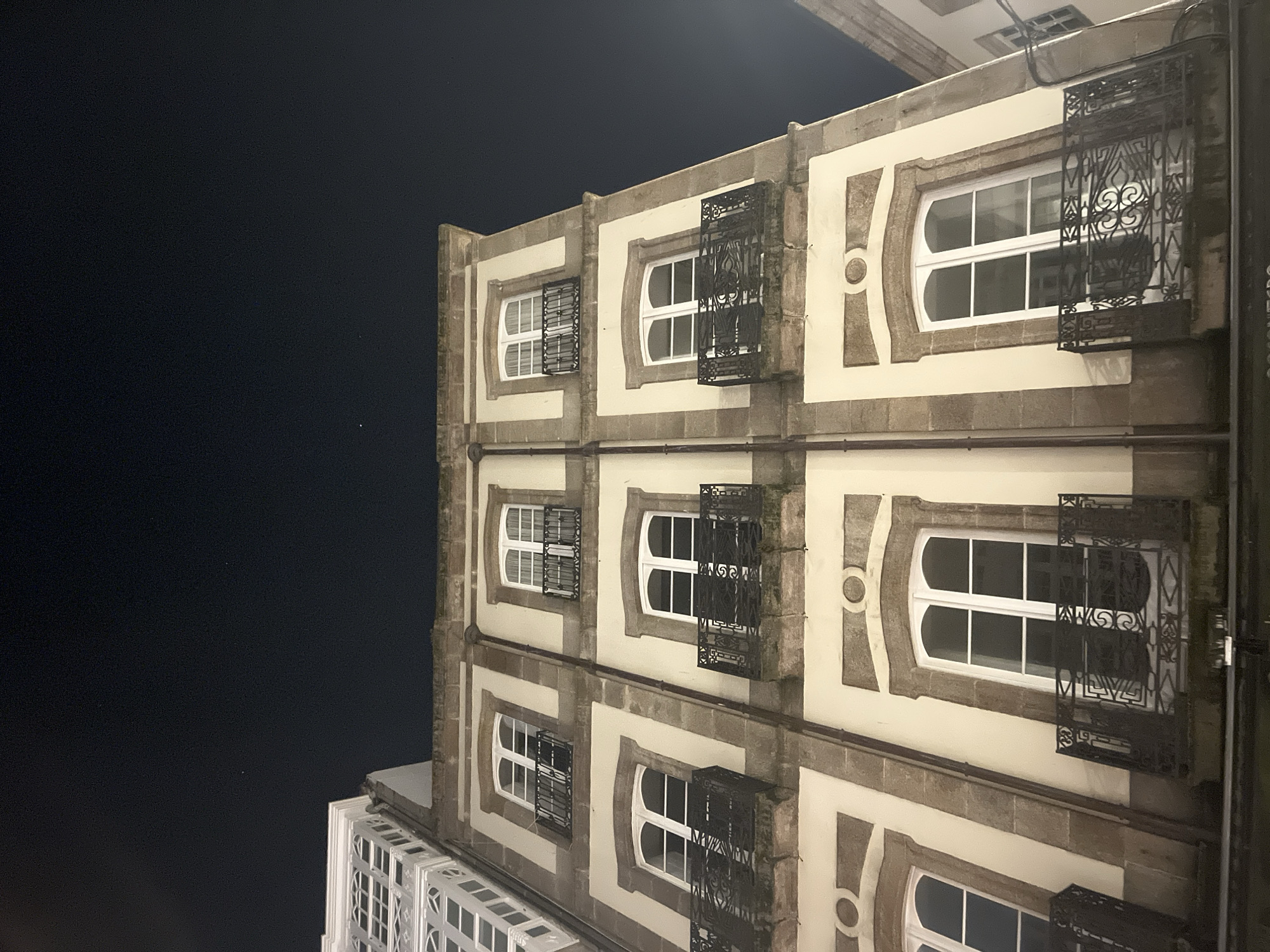 The image shows a four-story building in A Coruña, Spain, at night. The building features ornate windows with decorative ironwork grilles. The façade is a light color with stone accents. The sky is dark, indicating nighttime. Another building with white windows is visible on the bottom left of the image.