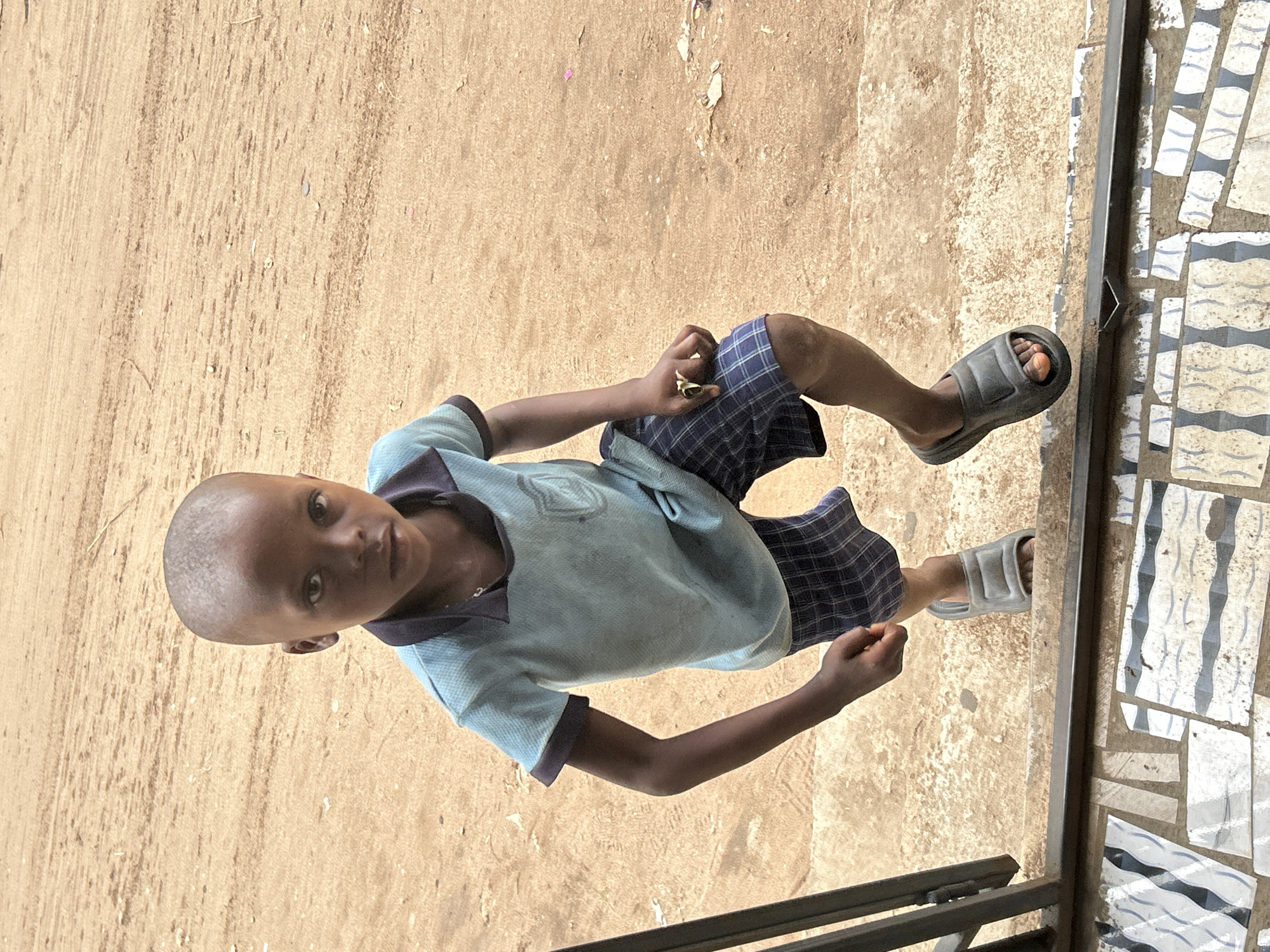 A young boy stands on a tiled surface adjacent to a dirt or concrete area, near a metal railing. He wears a light blue polo shirt, plaid shorts, and gray sandals. He is looking at the camera. The photo was taken in Ihiagwa, Nigeria.