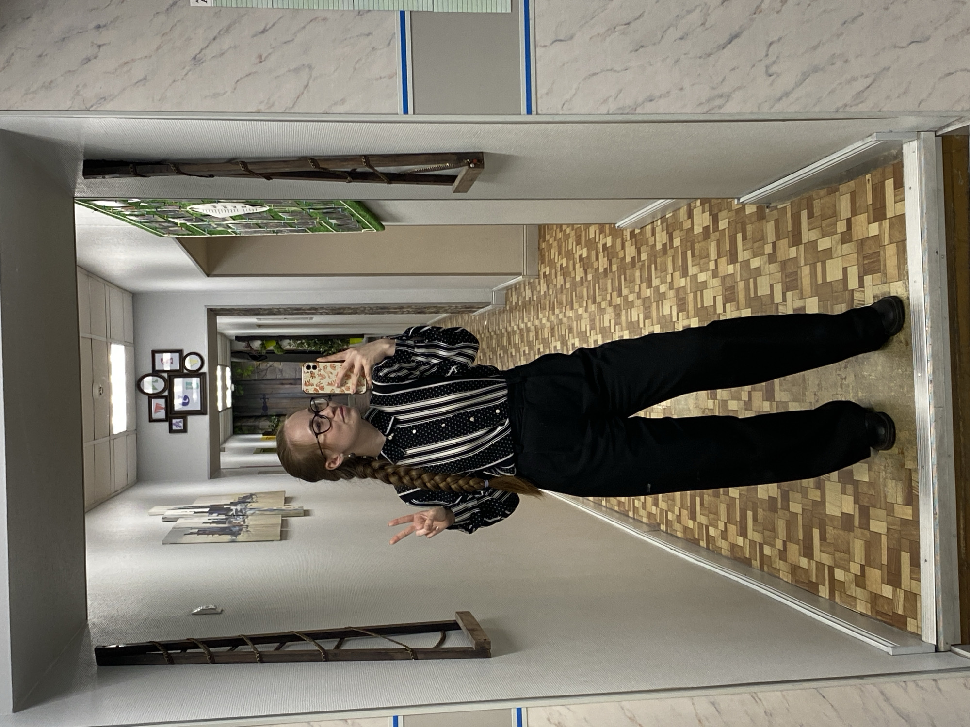 A mirror reflects a young woman in a hallway. The woman has red hair braided down her back, glasses, and is wearing a black and white striped shirt with black pants and shoes. She is holding a phone and making a peace sign. The hallway behind her has pictures on the wall, a wooden ladder, and a tiled floor. The ceiling is white with marble-like patterns. The location is not discernible from the image.