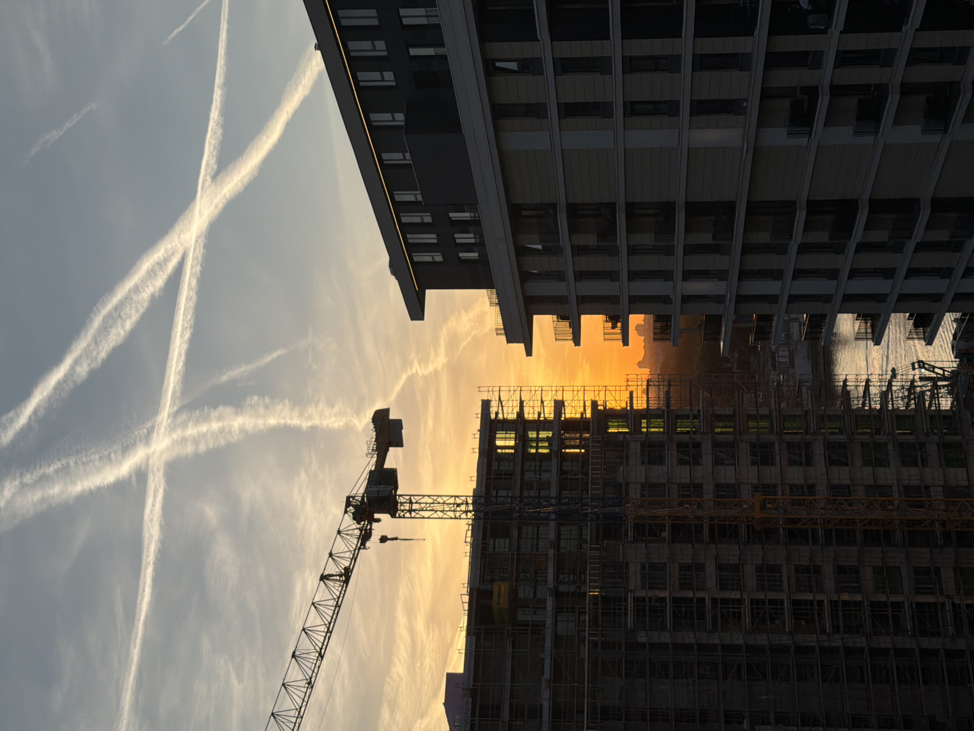 The image shows a view upwards between two tall buildings in Belgrade, Serbia. On the left, contrails are visible in the sky. In the center, a construction crane is silhouetted against the sky near a building under construction. The right side of the image features an occupied multi-story building. The sky has an orange glow from the setting sun.