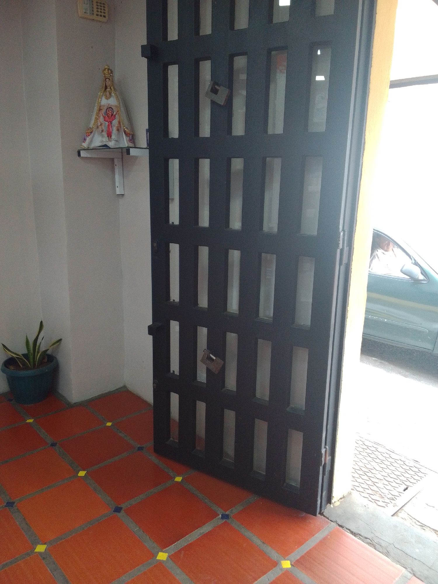 The image captures an outdoor scene in Juan Griego, Venezuela. In the foreground, a dark metal security door with glass inserts stands open, revealing a glimpse of a street and a person in a car. The door is secured with a padlock. To the left of the door, a pale grey wall displays a small religious figurine on a white shelf. Below the shelf, a potted plant with green and yellow striped leaves sits on an orange tiled floor. The tiles are arranged in a grid pattern with grey grout lines, interspersed with small yellow and blue squares. The overall impression is of an entryway with security measures and decorative elements.