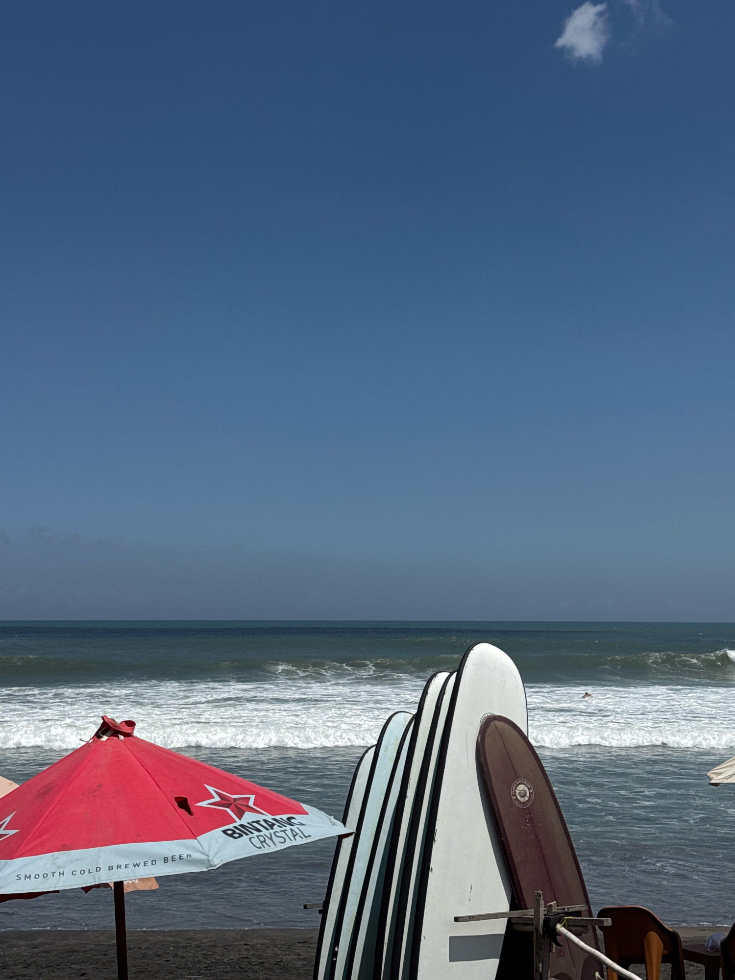 The image depicts a beach scene on a sunny day with a clear blue sky. In the foreground, a red umbrella with white text advertising "BINTANG CRYSTAL SMOOTH COLD BREWED BEER" stands on black sand. Beside the umbrella, several surfboards are propped up, leaning against each other. The surfboards are of various designs, with some featuring black and white stripes and others a solid brown color.
In the middle ground, the ocean stretches out with rolling waves. A person is visible in the distance, appearing to be a surfer in the water. Further back, the horizon is visible where the ocean meets the sky, with a hazy line suggesting land or clouds in the far distance. A single, small white cloud is present in the upper right portion of the sky. The overall atmosphere is one of a typical beach setting, likely a popular spot for surfing and relaxation. The location is identified as Unknown, Unknown.