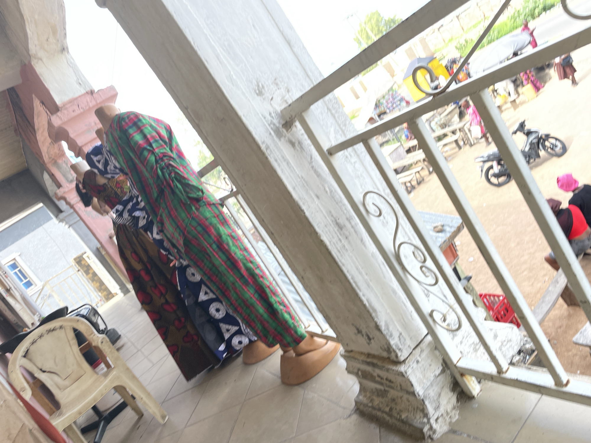 In the city of Jalingo, Nigeria, an outdoor scene unfolds. The image, taken from a slightly elevated perspective, is dominated by a white metal railing with decorative scrollwork, suggesting a porch or balcony. Below the railing, a section of tiled floor is visible, with a worn and partially damaged concrete pillar to the left.
Beyond the railing, the scene transitions to what appears to be an open area or courtyard. To the left, a red plastic crate sits on what looks like a wooden surface, surrounded by various objects, including a motorcycle with its front wheel visible. Further back, other items are scattered, some appearing to be furniture or building materials.
The focus shifts to the right of the image where two individuals are partially visible. One person, wearing a green and red plaid garment, is standing or leaning. Another person, wearing a blue and white patterned fabric, is also present, their interaction unclear. Behind them, a white plastic chair is overturned. The background reveals a building with visible window frames, and other structures in the distance, suggesting a residential or commercial area. The overall impression is of a somewhat cluttered outdoor space, possibly a marketplace or a residential backyard.