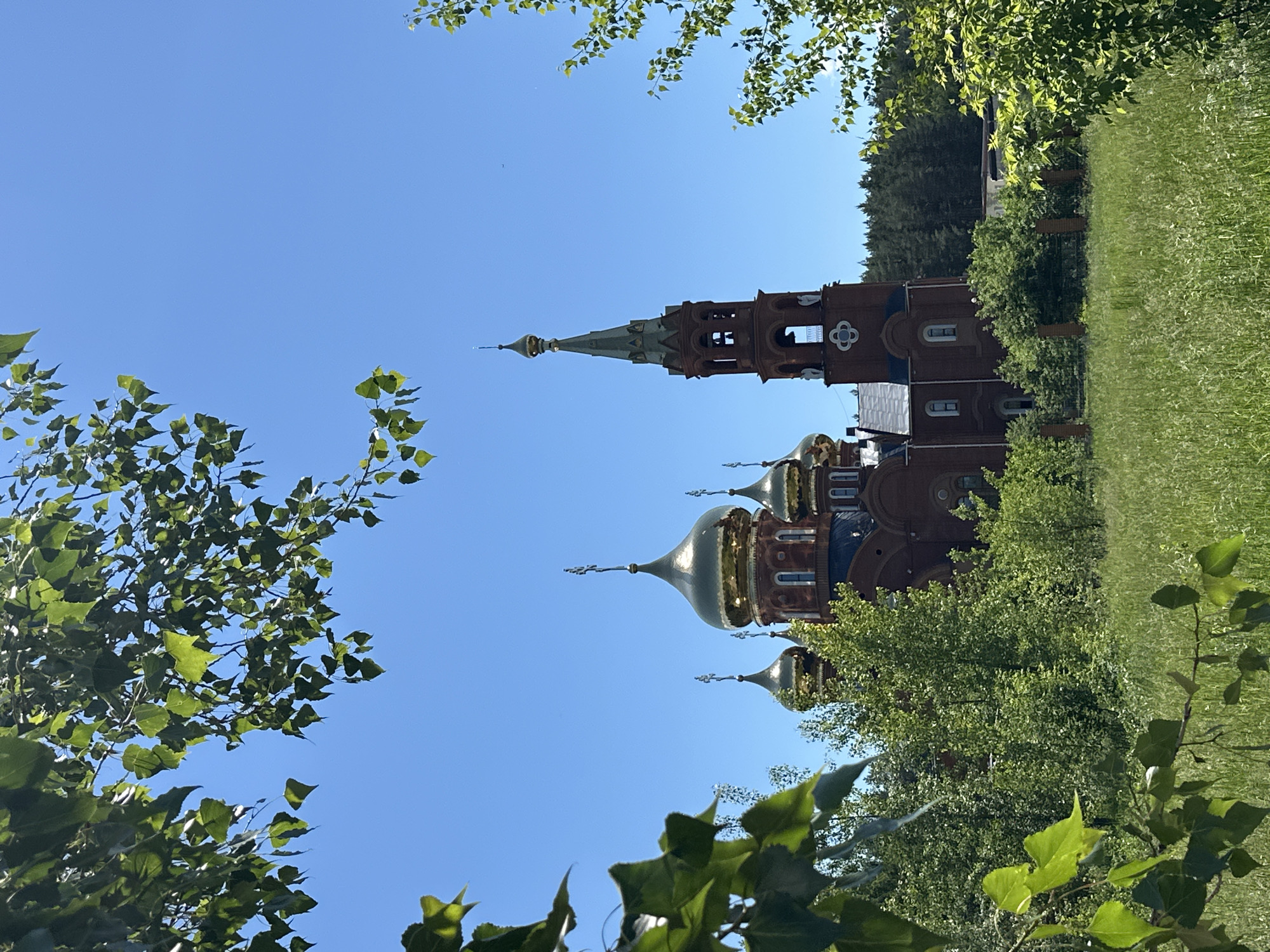 An Orthodox church with multiple golden domes and a tall bell tower stands amidst lush green trees and foliage under a clear blue sky. The church is constructed of red brick. In the foreground, out-of-focus green leaves and branches frame the scene. The church appears to be located in a somewhat rural or park-like setting, with a fence visible in the middle ground. The bell tower has arched openings and decorative elements. The overall impression is one of a serene and picturesque religious site. The church is likely the Holy Trinity Cathedral in Chaikovsky, Russia.