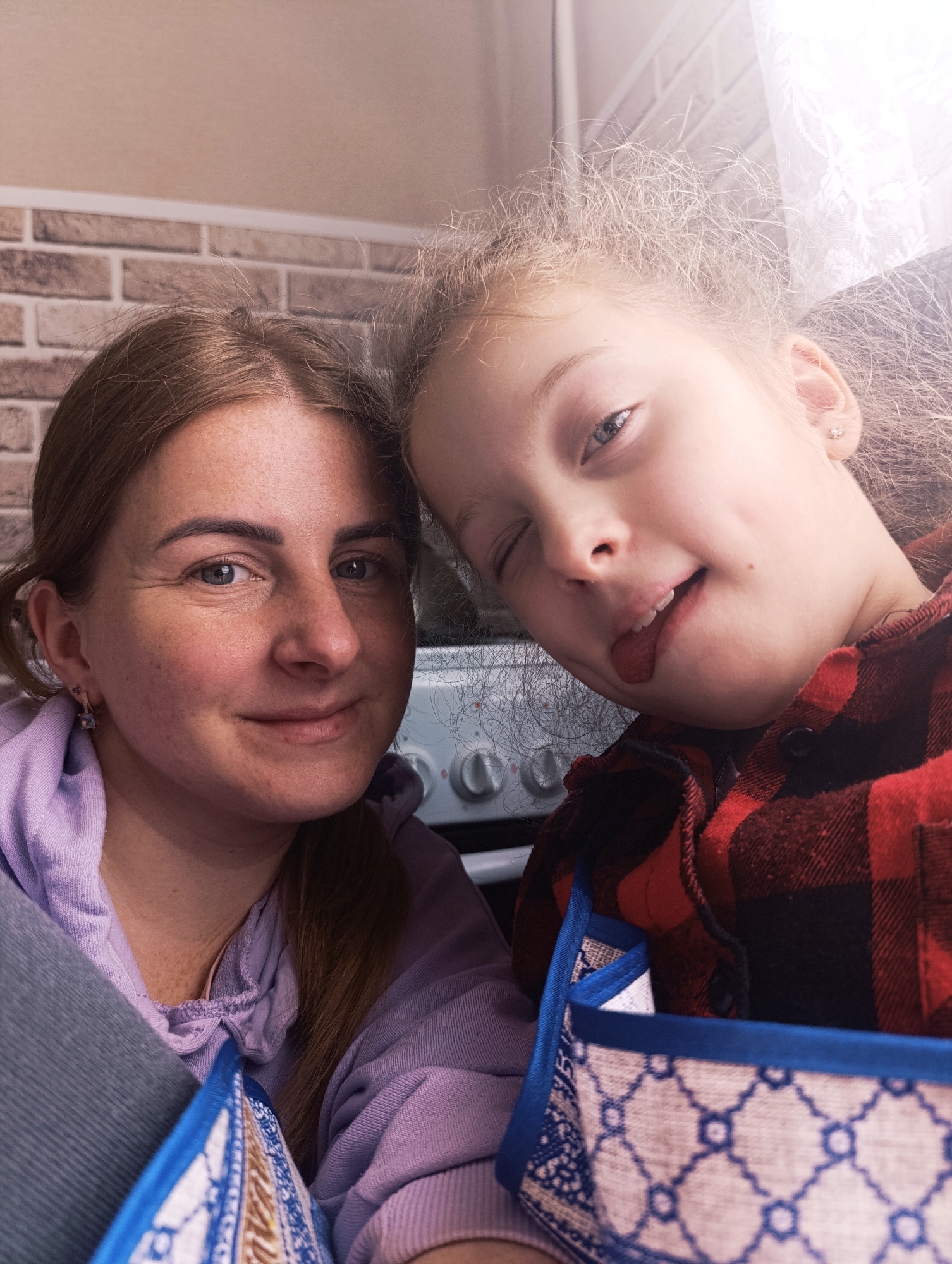 A medium shot captures a woman and a young girl in a close, indoor setting. The woman, on the left, has light skin, brown hair pulled back, and wears a light purple hooded sweatshirt. Her facial expression is a soft smile. The girl, positioned on the right, has light skin, blond curly hair, and wears a red and black plaid shirt. She is playfully sticking her tongue out and winking. Both are looking directly towards the camera.
The background includes a brick wall and a kitchen appliance, presumably an oven, with visible knobs. There's also what appears to be a patterned container or bag in the foreground. The environment is well-lit, with natural light coming from an unseen window.
There is no mention of a specific location, city, or state. The details suggest a casual, personal moment captured within a domestic setting.