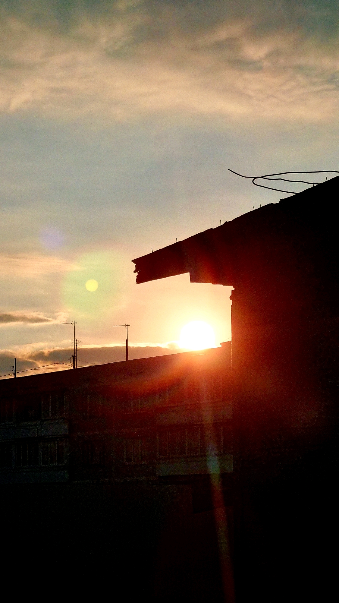 A silhouette of buildings and the sky at sunset. The sun is visible, low in the sky, and casting a warm glow. The sky is a mix of orange, yellow, and blue, with some clouds. The buildings are dark and featureless, with antennas on one of them. There are lens flares from the sun. The scene is of a city at sunset.