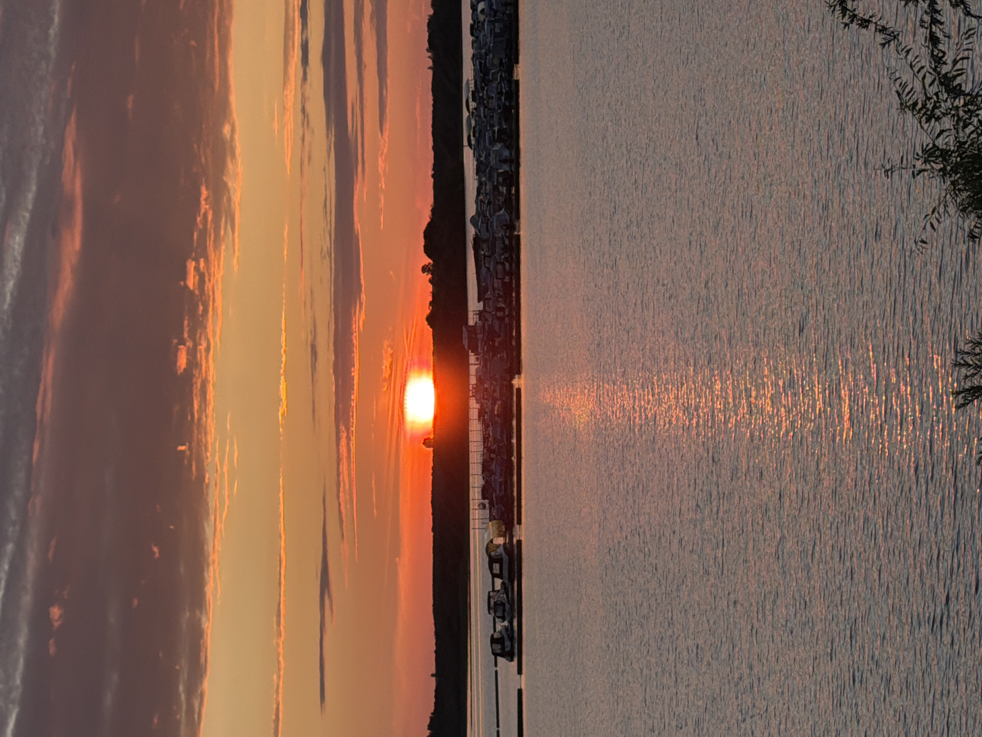 The image depicts a sunset over a body of water, likely a river or a lake, with a marina in the foreground. The sky is painted with vibrant hues of orange, yellow, and red, with dramatic clouds casting shadows. The sun, a brilliant orb, is setting on the horizon, casting a golden reflection on the rippling water.
The marina is filled with numerous boats docked side-by-side. The boats appear to be of various sizes and types, some covered with tarps. The shoreline behind the marina is lined with trees, silhouetted against the colorful sky. The overall scene is tranquil and serene, showcasing the beauty of nature at dusk.
While the image is evocative and visually stunning, it does not contain enough contextual information to definitively place it in Solikamsk, Russia, or any other specific city. The general landscape of a marina with a body of water and treelined shores is common to many locations. To provide a precise location, additional details such as landmarks, specific boat types, or signage would be required.
