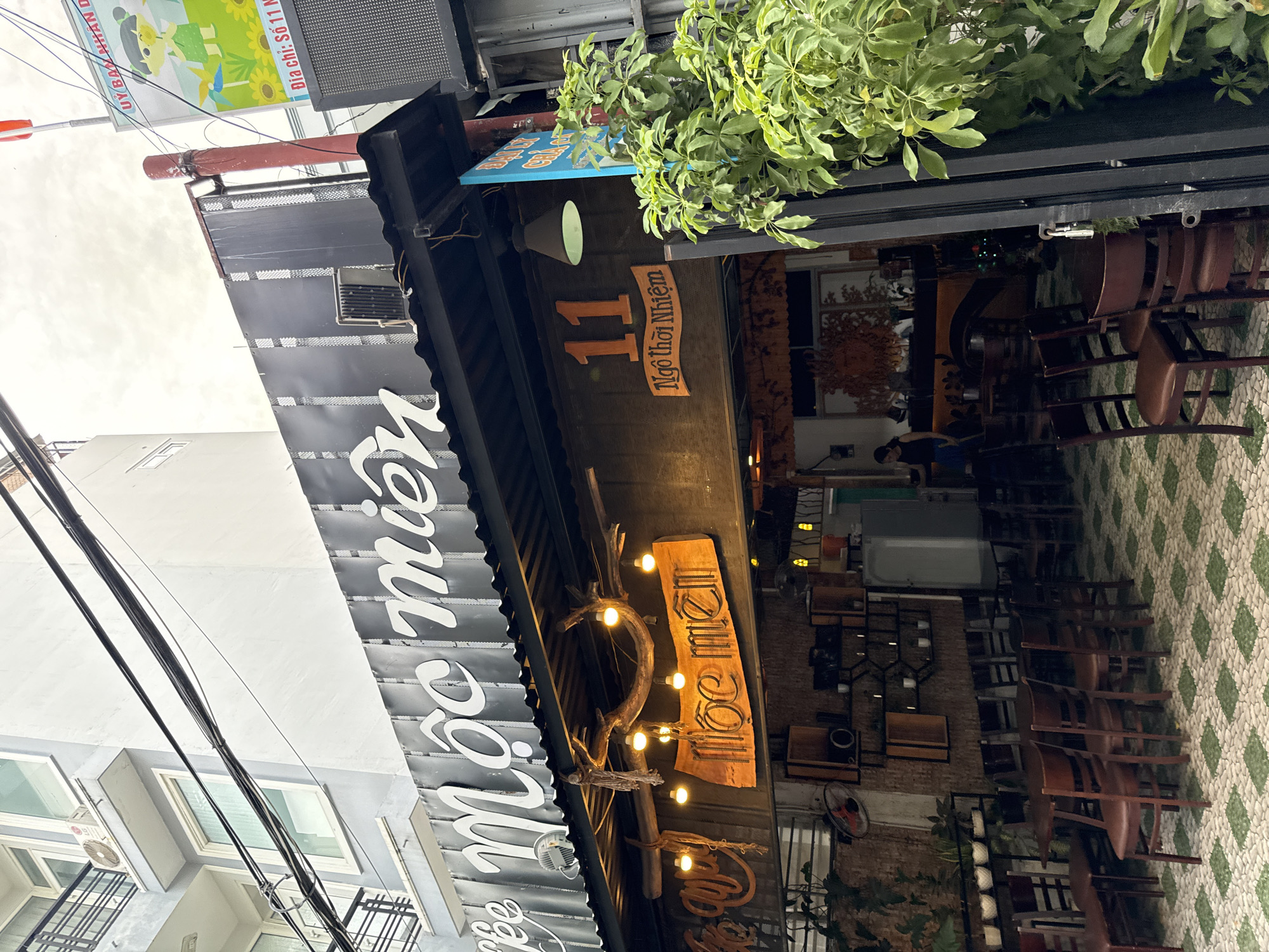 The image shows the exterior of a cafe named "Mộc Miên" located at 11 Ngô Thời Nhiệm street in Nha Trang, Vietnam. The cafe has a rustic design with wooden elements. Above the entrance, the cafe's name is written in large white script on a black background. Below this, a wooden sign displays the cafe's name again, along with the street number "11" and the street name "Ngô Thời Nhiệm." String lights hang from a wooden beam above the entrance, illuminating the seating area. The cafe appears to be open, with several tables and chairs visible inside and on the patio. A person is standing near the counter inside the cafe. A vibrant green plant is growing in front of the cafe on the right side of the image. A light pole with a red vertical stripe is visible to the right of the cafe. A billboard with a cartoon character and text is mounted on the pole.