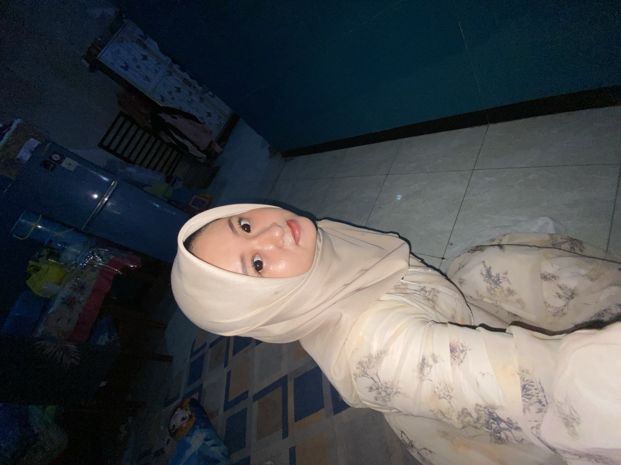 A person wearing a hijab and patterned dress is visible in an indoor setting. Behind the person are various objects including a refrigerator, crib, and other items. The person appears to be the focal point, while the other objects serve as background elements in an interior. The location is likely Gelam, Indonesia, based on the given information.