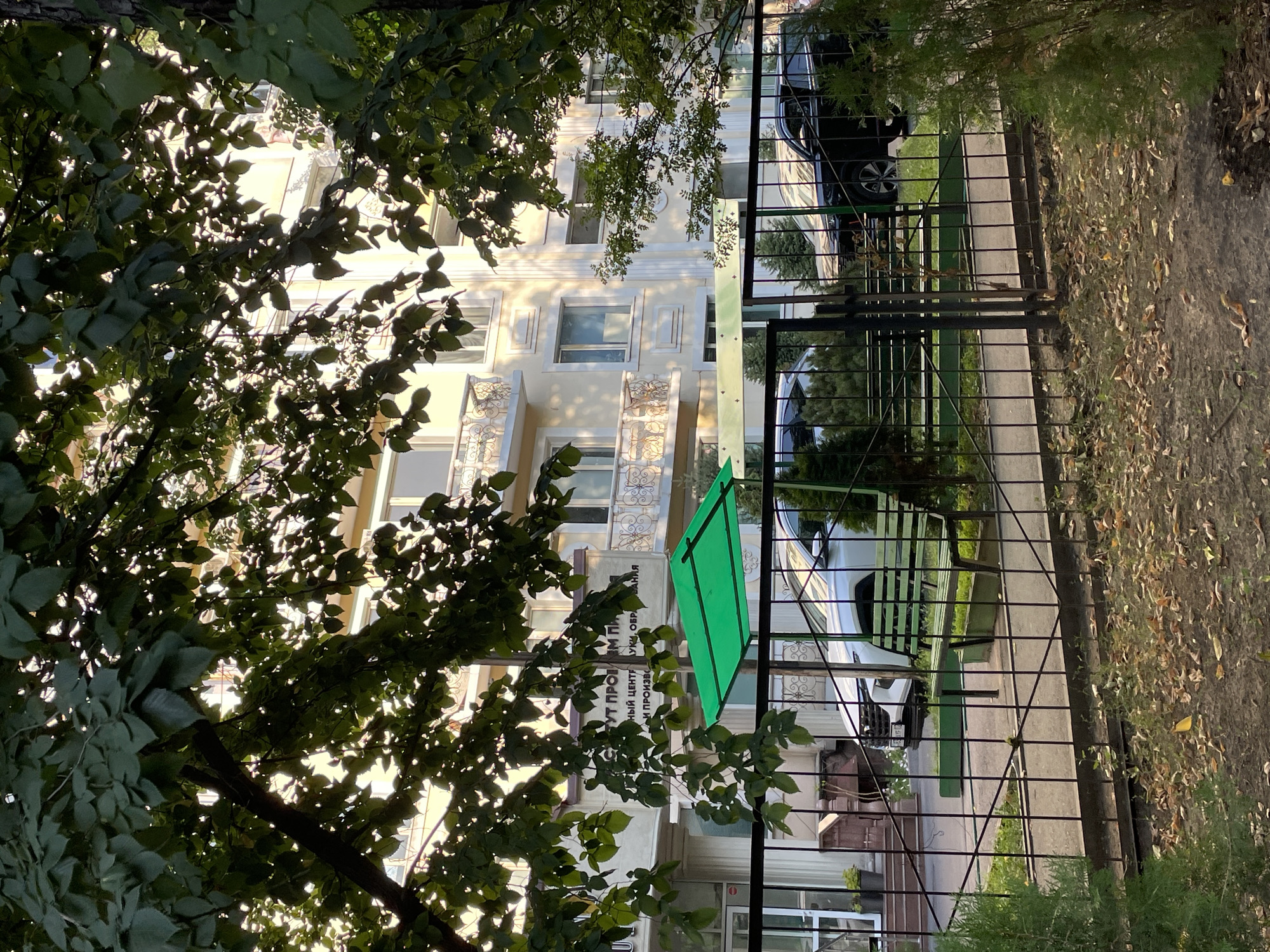 The image depicts the facade of a light-colored building in Almaty, Kazakhstan, partially obscured by lush green foliage. The building, possibly a medical or research facility given the partial view of signage that reads "СТУТ ПРОБЛЕМ ПЛ" and "НЫЙ ЦЕНТР УКИ, ОБРАЗОВАНИЯ И ПРОИЗВО", features balconies with decorative railings on its upper floors. In front of the building, a black metal fence encloses a green bench and a green awning. Two cars are visible parked behind the fence; a white SUV is on the left, and a black SUV is on the right. The ground in the foreground is covered with dirt and fallen leaves, with some sparse green plants. The lighting suggests it is either early morning or late afternoon.