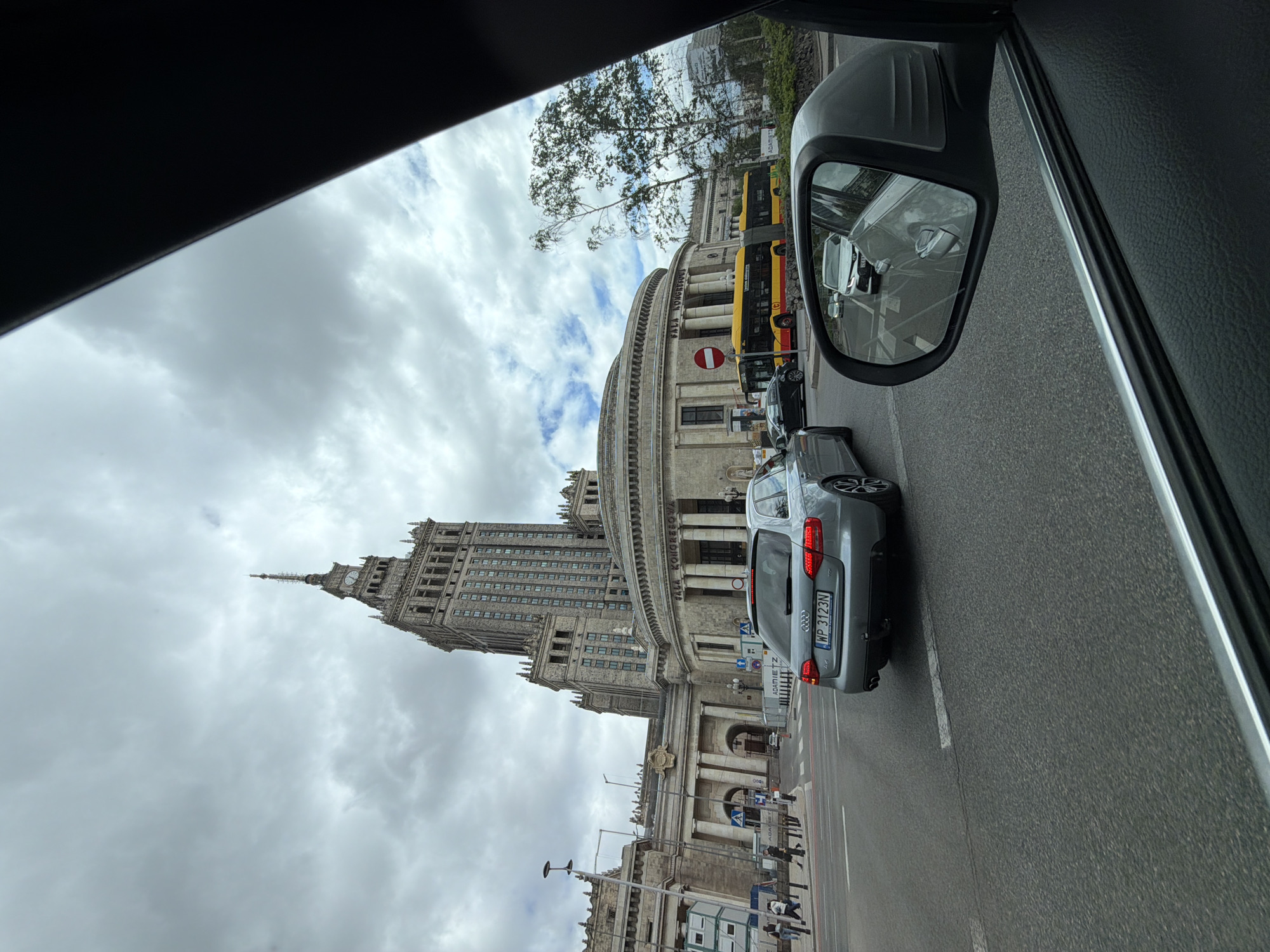 From inside a vehicle, the Palace of Culture and Science in Warsaw, Poland is seen on the left. A silver Audi A4 with license plate WP 3123N is in the adjacent lane. A yellow bus is visible in the side mirror. Pedestrians are walking on the sidewalk in front of the building. The sky is overcast.