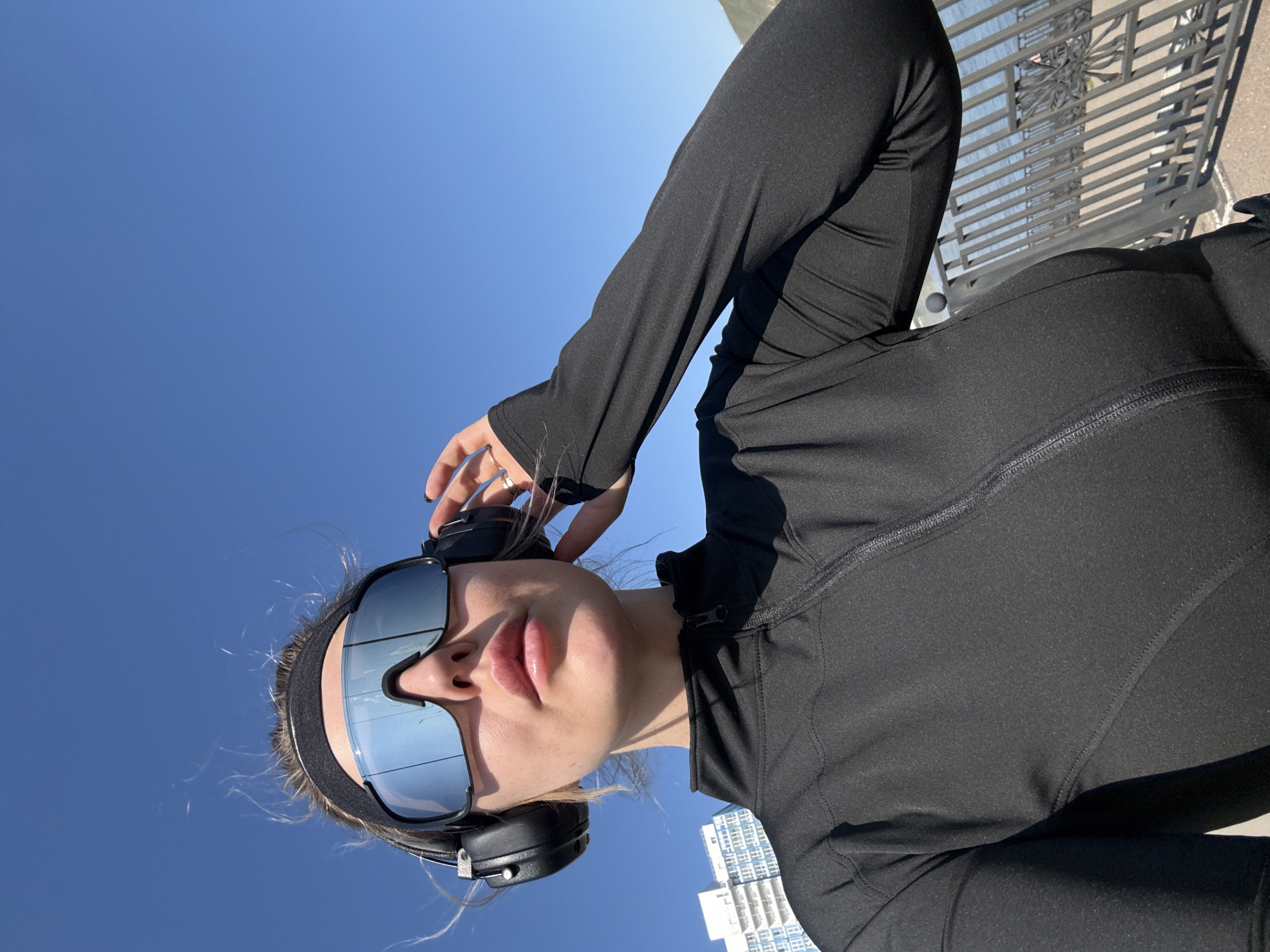 A low-angle photograph depicts a person wearing sunglasses, headphones, and a black long-sleeved jacket, posing against a clear blue sky. The person's right hand is near their head, appearing to adjust the headphones or hair. The image shows the city skyline, with a white high-rise building visible in the background. The location appears to be in Krasnoyarsk, Russia.