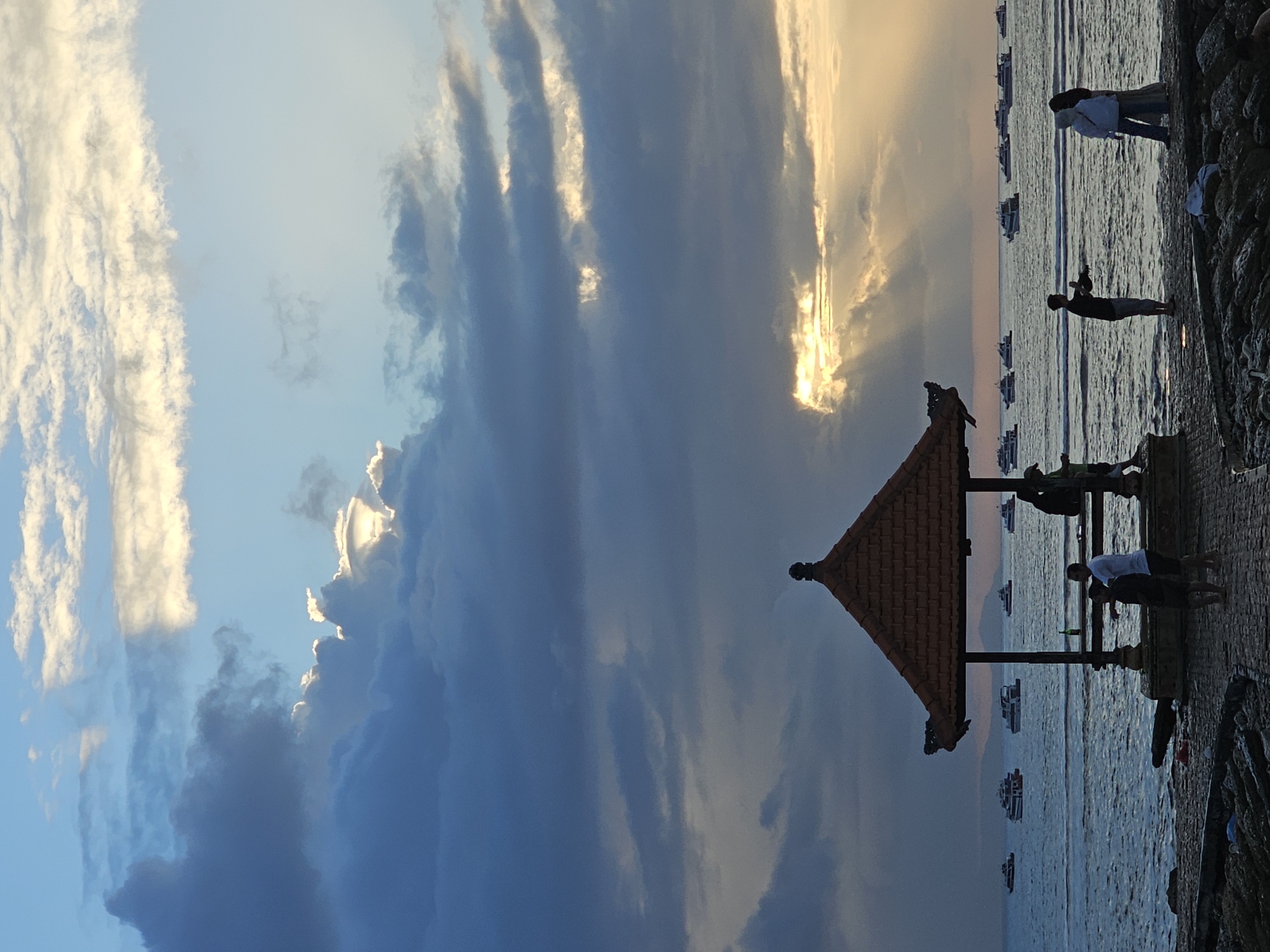 A vertical, silhouette-heavy, medium shot captures a tranquil sunset scene at Sanur Beach, Denpasar, Indonesia. The sky dominates the upper two-thirds of the frame, a dramatic canvas of deep blues and grays from turbulent clouds, pierced by brilliant rays of golden sunlight breaking through in the mid-to-upper right. These rays create a divine, ethereal glow, illuminating portions of the clouds and casting subtle highlights. The lower third of the image features the calm, dark blue ocean, dotted with numerous white boats of varying sizes anchored offshore. A traditional Balinese-style gazebo with an orange tiled roof stands prominently in the foreground, slightly to the left of center. Several silhouetted figures are scattered around the gazebo and the stone-paved walkway leading to it. On the walkway in front of the gazebo, two men, one in a white shirt and the other in a dark shirt, are visible. To the right of the gazebo, a man in a dark shirt is seen taking a photo or video with his phone. Further to the right, a woman in a hijab and a light-colored top and skirt, and another woman in a dark headscarf and light clothing, are conversing. The foreground is composed of rough, dark rocks and a stone pathway, suggesting a breakwater or pier. The overall atmosphere is one of peaceful evening serenity, with the dramatic sky and the silhouettes of people and structures creating a striking contrast.