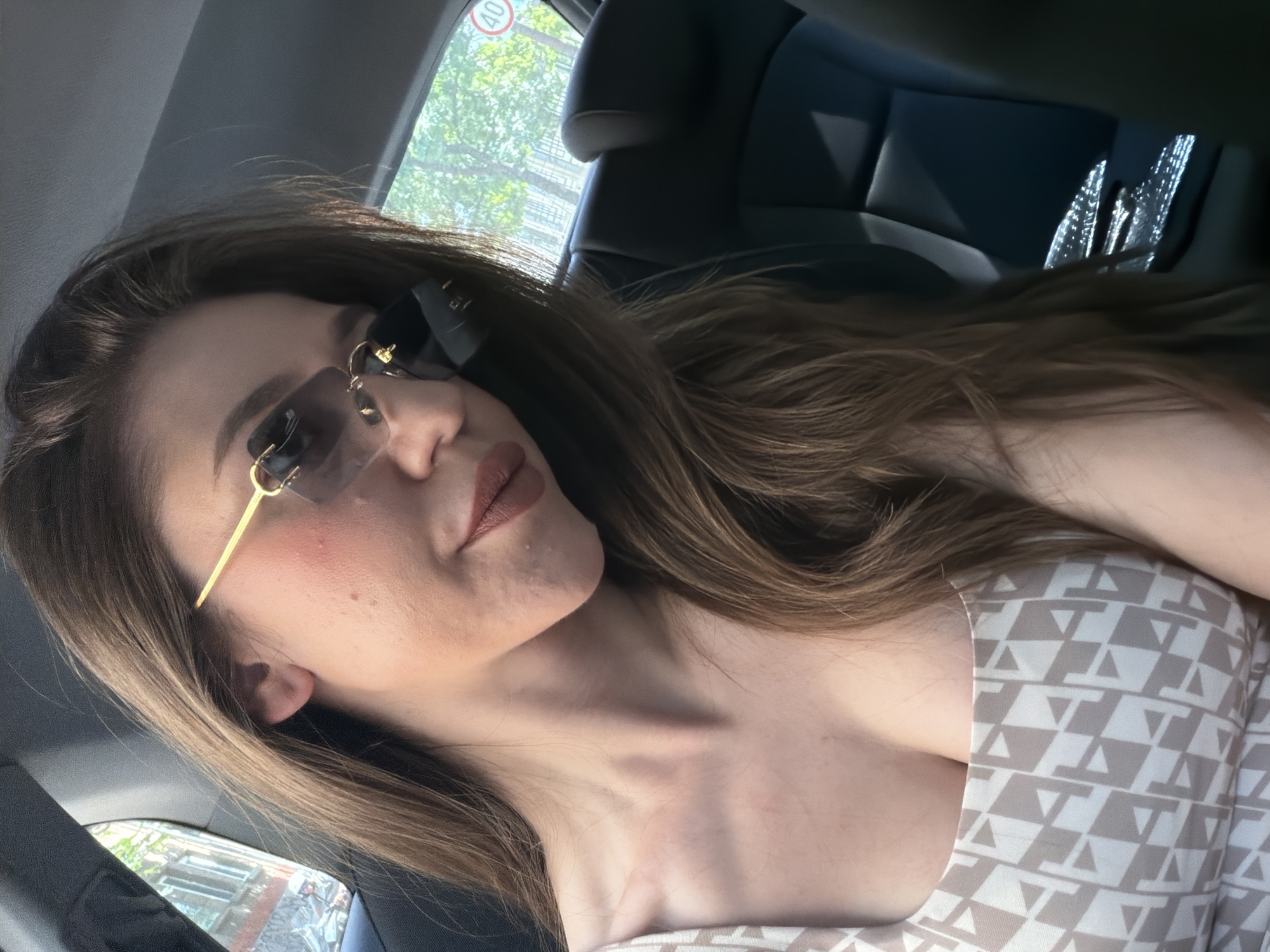 A woman with long brown hair and sunglasses is sitting in the back seat of a car. She is wearing a cream-colored dress with a geometric pattern. Her hair is partially covering her face and shoulders. The interior of the car is visible, with dark leather seats and a headrest. Outside the window, trees and buildings can be seen, suggesting an urban environment. A speed limit sign with "40" is visible on a pole outside the car. The lighting indicates it is daytime. The overall scene depicts a woman traveling in a car, likely in a city. Sofia, Bulgaria has many trees and urban settings, making this a plausible location.