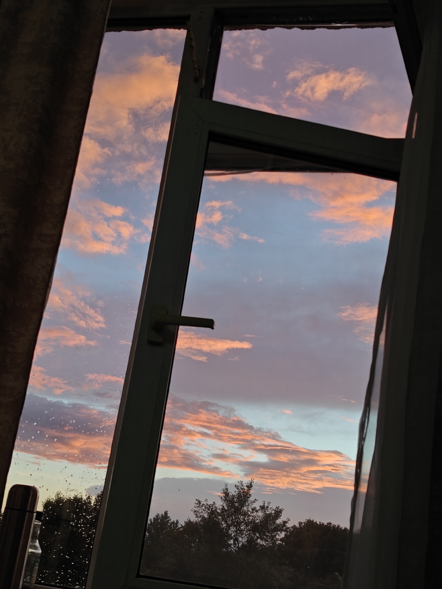 The media file is a photograph taken from inside a building, looking out through a window at a sunset sky. The window has a white frame and a metal handle. There are curtains on the left and right sides of the window, partially obscuring the view. The sky is filled with clouds that are illuminated by the setting sun, displaying a gradient of colors from pale pink and orange to light blue and purple. The clouds appear textured and somewhat wispy.
Through the lower part of the window, the silhouettes of trees are visible against the colorful sky. The trees appear dense and dark, suggesting they are not directly illuminated by the sunset. On the left pane of the window, water droplets are scattered across the glass, indicating that it might be raining or has recently rained, adding a layer of texture to the scene. A thermos and a bottle are visible on a surface in the lower left corner, inside the room.
The time of day is clearly sunset, evidenced by the dramatic colors in the sky. The presence of clouds and the light suggests a possibly overcast or partly cloudy day. The emotional tone evoked is serene and contemplative, with the beauty of the sunset contrasting with the indoor perspective and the hint of weather. There is no visible text in the image. The location context is Pushkino, Russia.