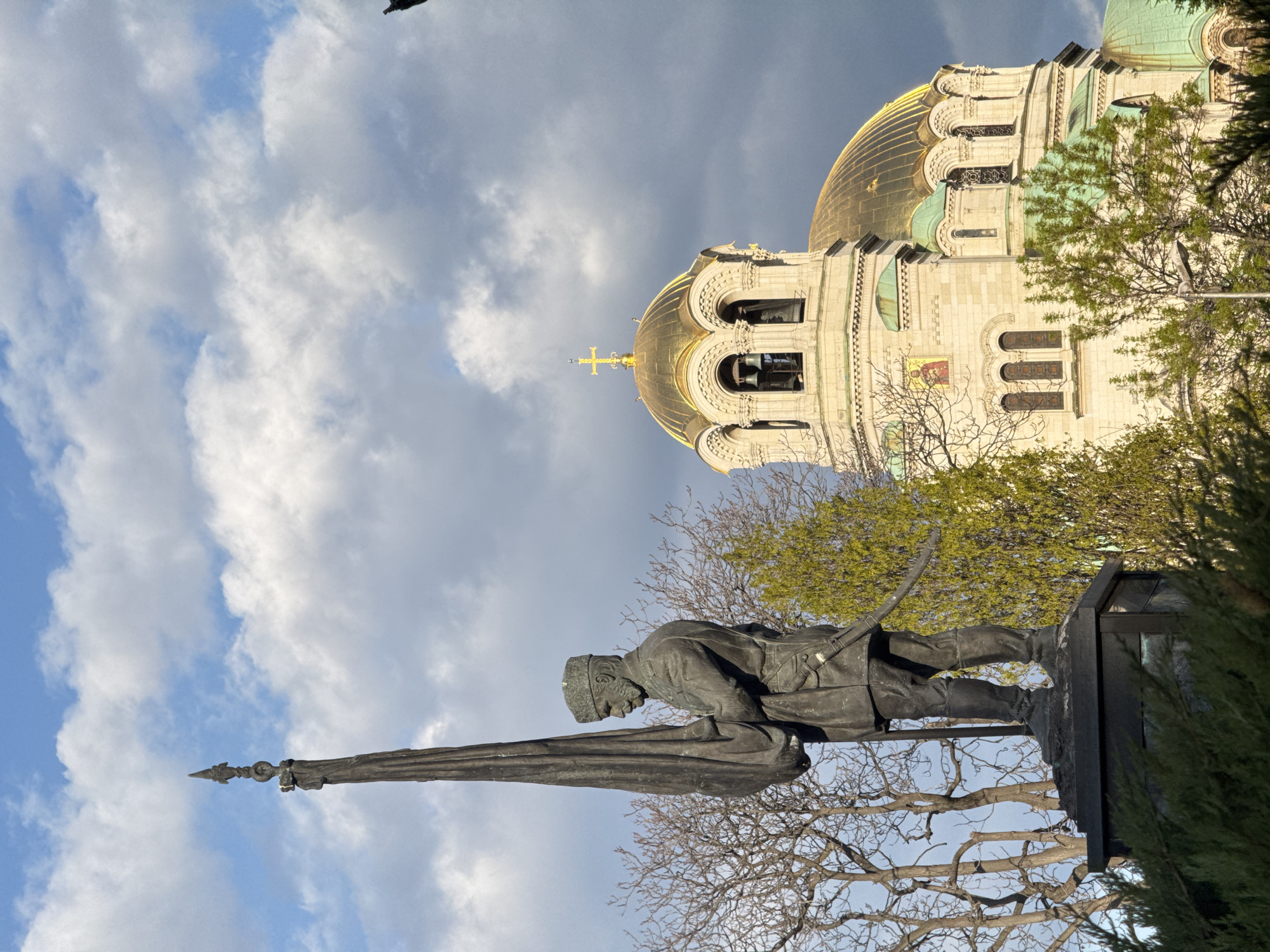The image, correctly oriented, displays a bronze statue of a soldier in the foreground with a large, majestic cathedral in the background, set against a partly cloudy sky. The scene is located in Sofia, Bulgaria, and prominently features the Alexander Nevsky Cathedral.
The statue, positioned on a dark stone pedestal, depicts a male soldier dressed in a military uniform with a cap. He holds a tall, dark standard or flag upright. Bare tree branches are visible behind and to the sides of the statue, suggesting late winter or early spring before deciduous trees have fully leafed out.
Dominating the background is the grand Alexander Nevsky Cathedral, an Orthodox structure characterized by its distinctive golden domes. One prominent dome features a cross at its apex, while another includes a bell tower section with visible bells. The cathedral's facade is constructed from light-colored stone and exhibits intricate architectural details, such as arched windows and various decorative elements. Some sections of the roof and smaller domes are finished in a greenish-teal color. Lush green foliage from other trees is visible around the cathedral, contrasting with the more barren trees closer to the statue. A small, colorful icon or painting is visible on one of the lower walls of the cathedral.
The sky is a mix of bright blue patches and scattered white to light grey clouds, indicating a clear but not entirely cloudless day. The natural lighting suggests daytime, possibly mid-morning or afternoon. No specific activity or event is taking place; the image captures the architectural and monumental presence of these significant structures. There is no discernible text visible on either the statue or the cathedral.