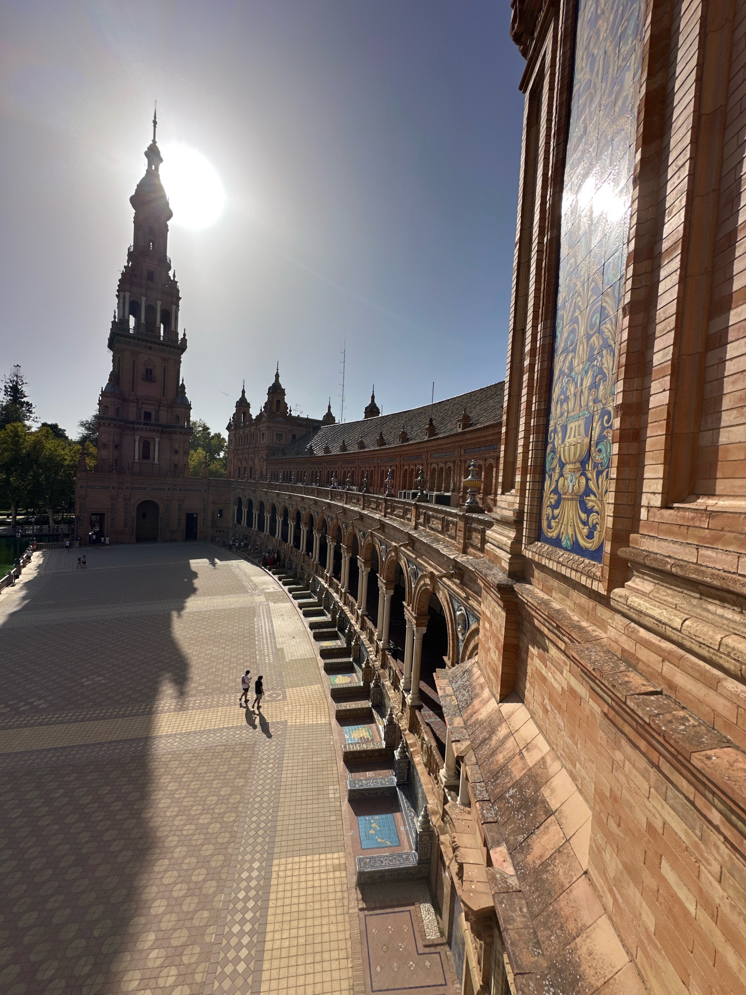 This image captures the Plaza de España in Seville, Spain. The scene is dominated by the sun, which is positioned in the upper left quadrant of the sky, casting long shadows across the tiled plaza. In the foreground, the right side of the frame features a close-up of a brick building with decorative blue and gold tiles. The building curves away from the viewer, revealing a colonnaded walkway with arches. Two figures, appearing to be young adults, walk across the sunlit plaza in the middle ground. Behind them, the plaza extends towards a large, ornate tower and the continuation of the curved building complex. Trees are visible in the background on the left side. The sky is a clear, light blue, with some hazy atmospheric effects from the bright sun. The overall impression is one of architectural grandeur bathed in the strong light of a sunny day.