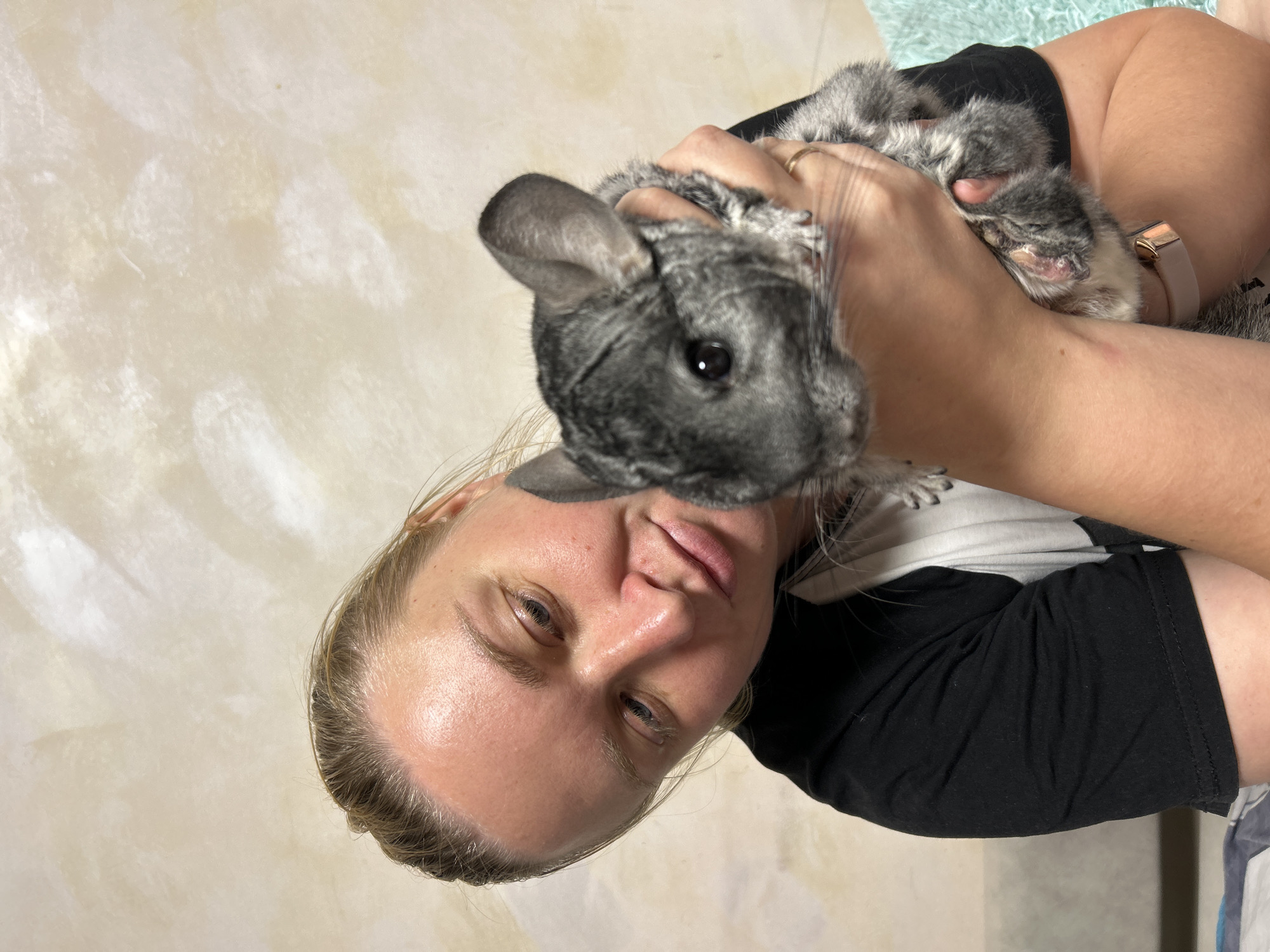 A woman with blonde hair pulled back is holding a chinchilla. The woman is wearing a black t-shirt with a white graphic. The chinchilla is gray with darker markings. The background appears to be a wall with textured paint. The scene is indoors.