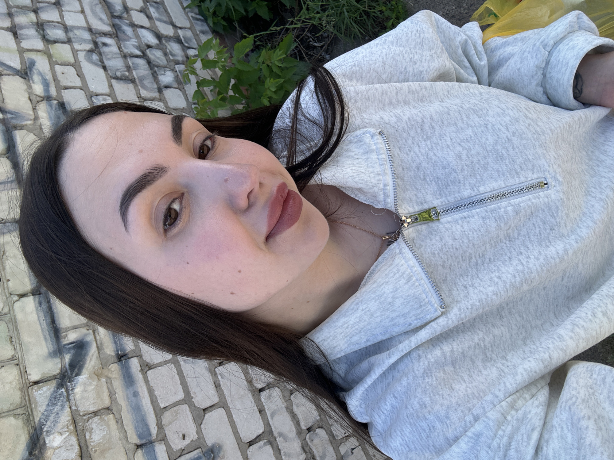 A young woman with dark hair and brown eyes is pictured outdoors. She wears a light gray zip-up hoodie. The background includes a brick pavement, green plants, and a yellow plastic bag. The location is not discernible as Фоки, Russia.