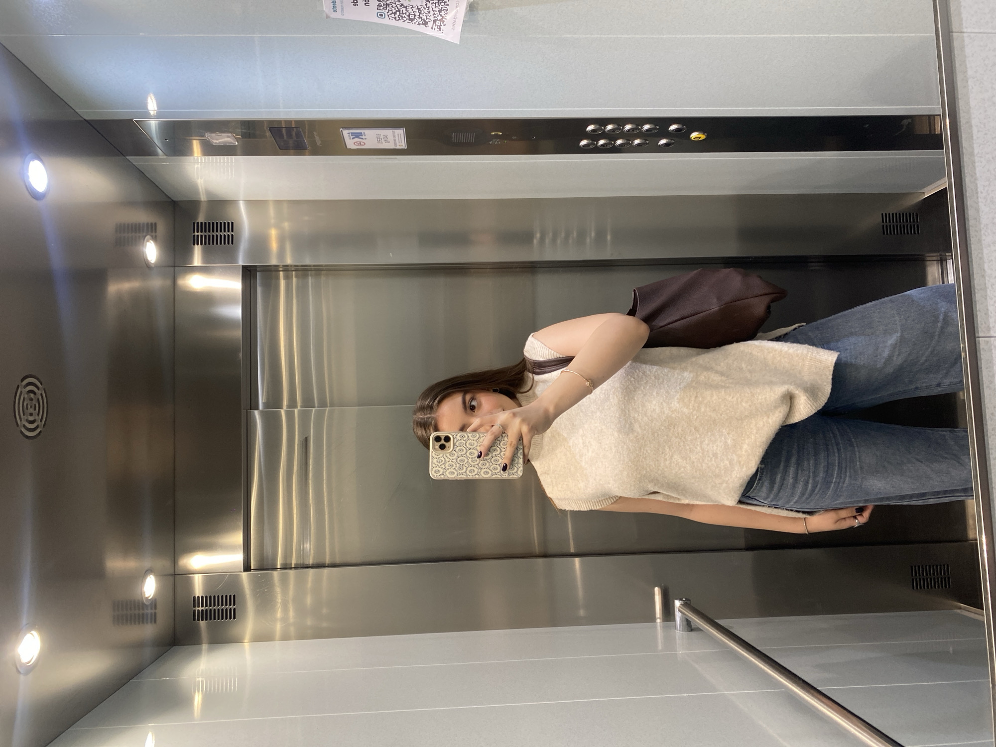 A woman is taking a mirror selfie inside of an elevator. She is wearing jeans and a sweater. She is holding a smartphone with a decorated case in front of her face. A brown bag is slung over her shoulder. The elevator is made of stainless steel and has lights on the left side and control buttons on the back wall. A handrail is located on the right wall. A QR code is visible in the top left corner of the elevator.