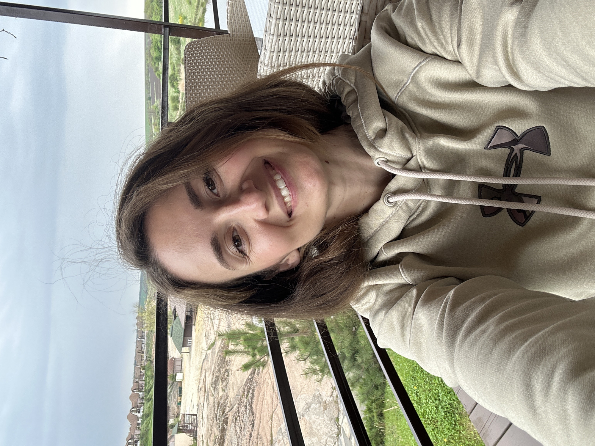 A woman is sitting in a wicker chair on a balcony overlooking a residential area of Zakharivka, Ukraine. She is wearing a light tan Under Armour hooded sweatshirt. The view from the balcony shows houses with brown rooftops and grassy areas and trees between buildings. The sky is overcast. The woman is looking at the camera and smiling. The balcony has dark metal railings.