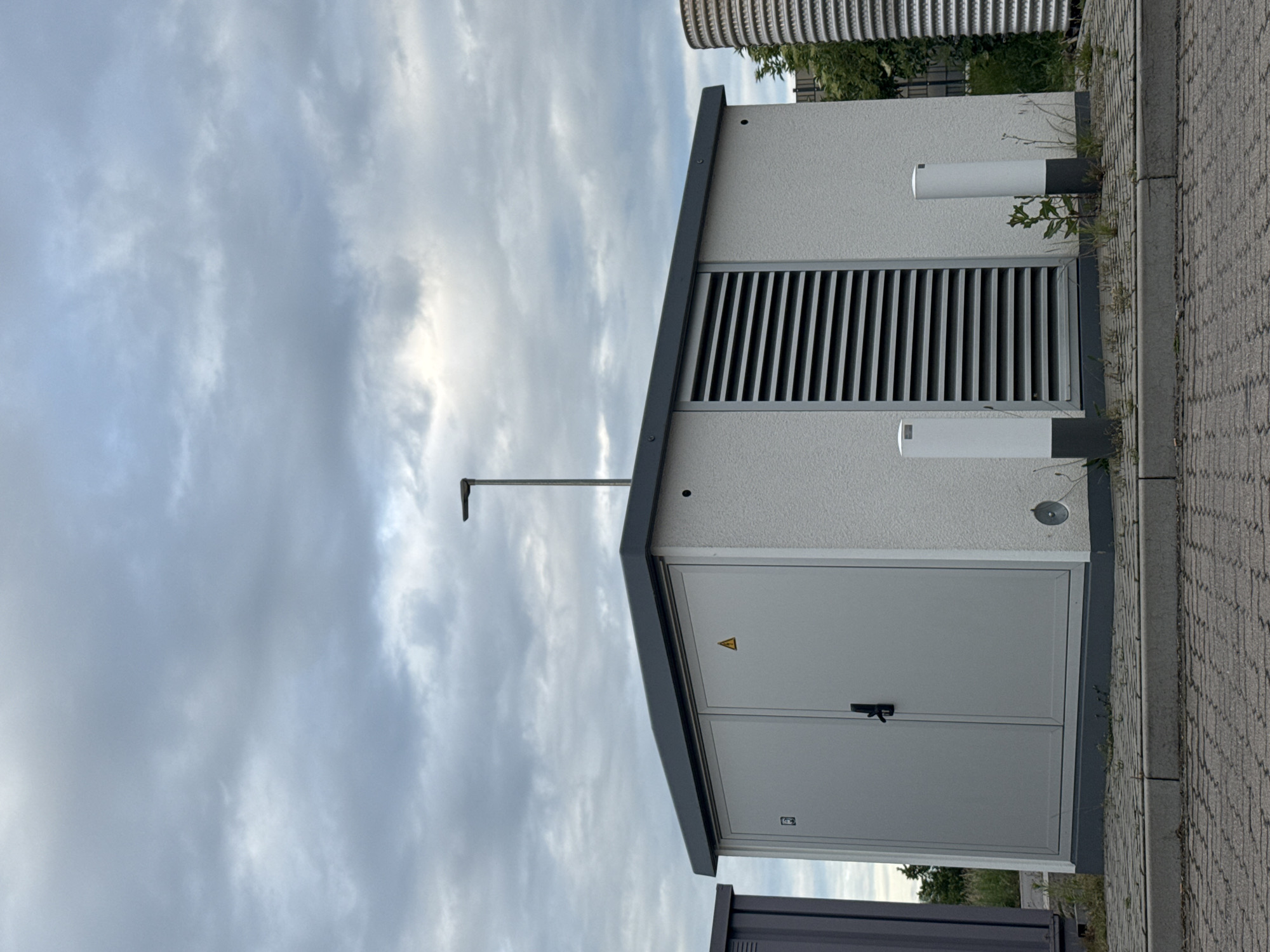 An outdoor medium shot shows a white, rectangular electrical transformer box with a dark gray flat roof. The box has double doors on the left side with a black handle and a yellow warning triangle symbol. To the right of the doors, a large vent with horizontal slats is visible. Two white cylindrical bollards with dark gray bases stand in front of the box. A light pole extends vertically from the top of the box. The ground in front of the box is paved with gray interlocking bricks and bordered by a gray concrete curb. In the background, a dark gray metal structure is partially visible on the left, and a ribbed, cylindrical object is on the right. Trees and vegetation are present behind the box. The sky is filled with gray, cloudy skies.