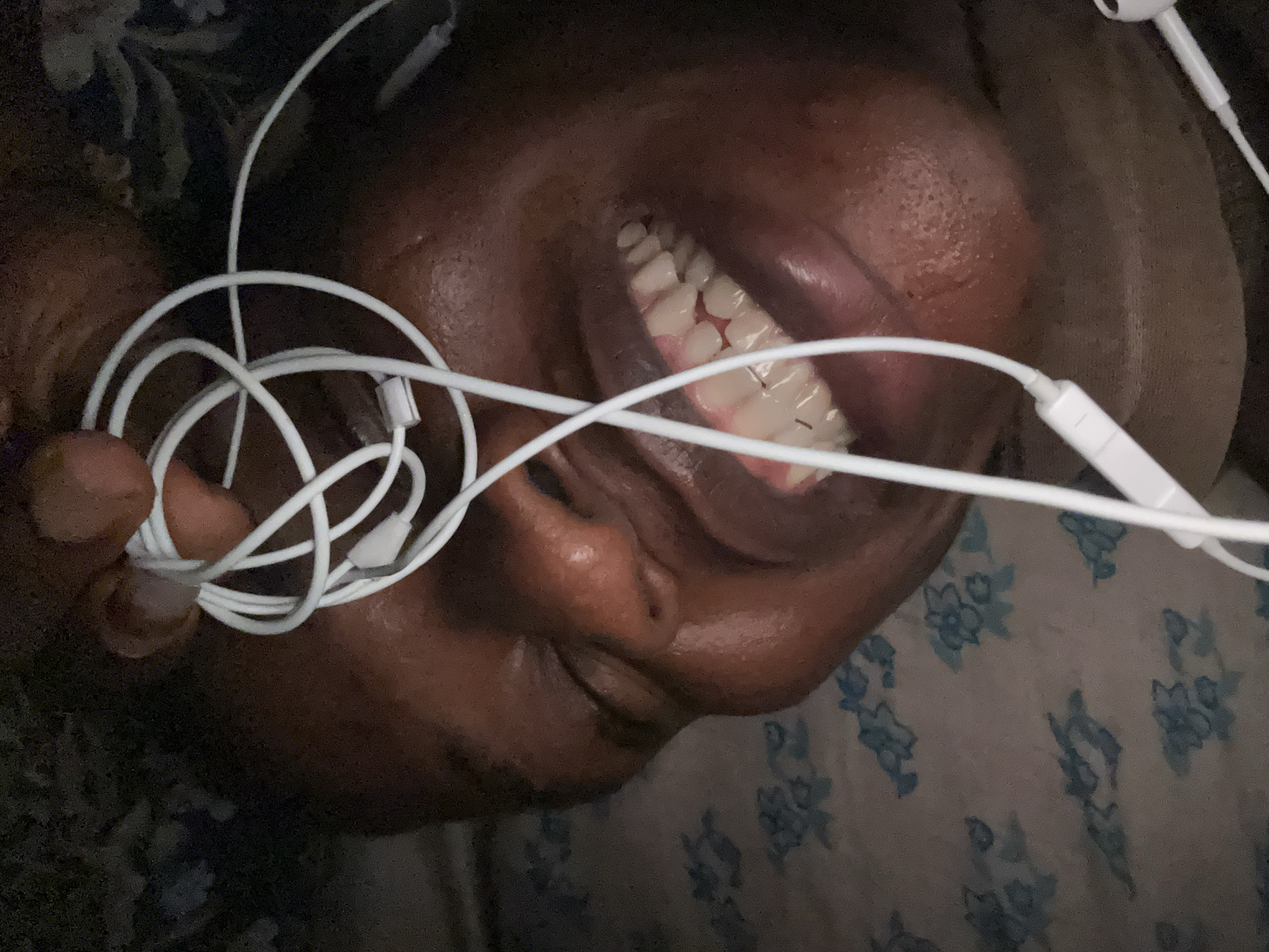 A close-up, upside-down photograph shows a person with dark skin smiling. White earbuds are in their ears and tangled in their hand. The person is lying on a surface with a light-colored fabric covered in blue floral patterns. The location and city are not identifiable.