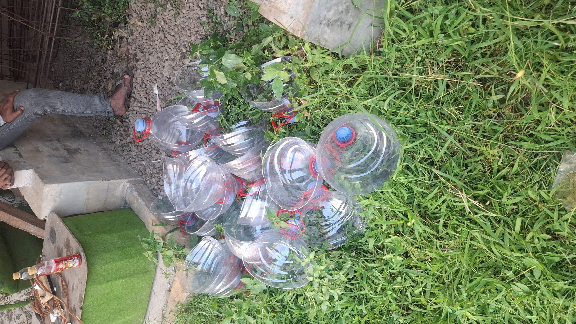 In Rahong, Indonesia, a collection of empty plastic bottles, some with red caps, lies scattered on a patch of grass and gravel. In the background, a person in gray jeans and sandals is partially visible, sitting near a concrete structure and a green wooden box. Several electrical wires and two small bottles, one red and one yellow, are on top of the green box. The scene appears to be outdoors, possibly in a yard or open area.