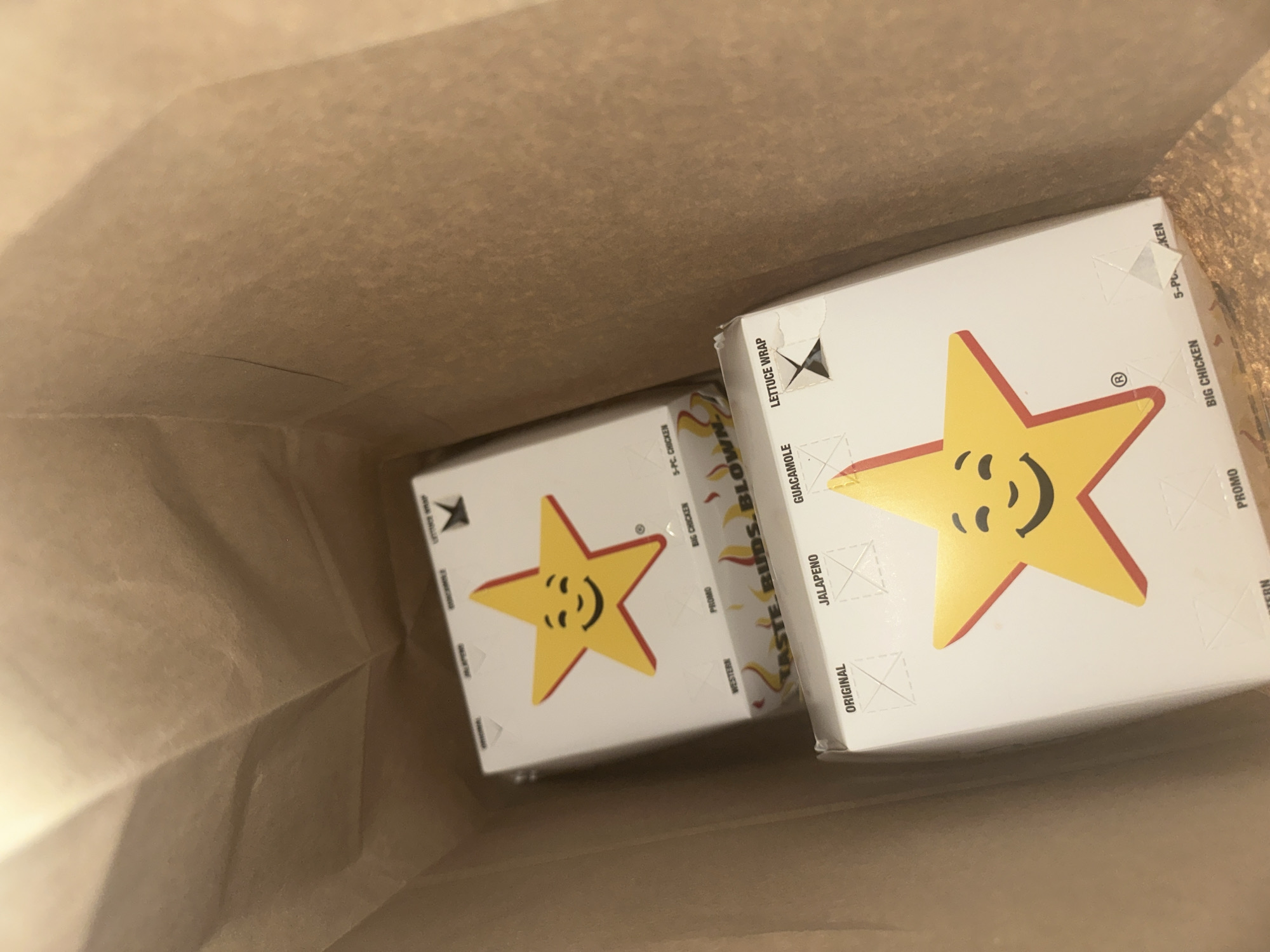 The image depicts two white cardboard boxes inside a brown paper bag. The boxes are printed with a yellow star logo outlined in red. The stars have a smiling face. Text printed on the boxes includes "WESTERN", "PROMO", "BIG CHICKEN", "5-PC. CHICKEN", "TASTE. BUDS. BLOWN", "ORIGINAL", "JALAPENO", "GUACAMOLE", and "LETTUCE WRAP". The image does not provide information about the location in San Bernardino Tlaxcalancingo, Mexico, people, actions, interactions, or the environment.