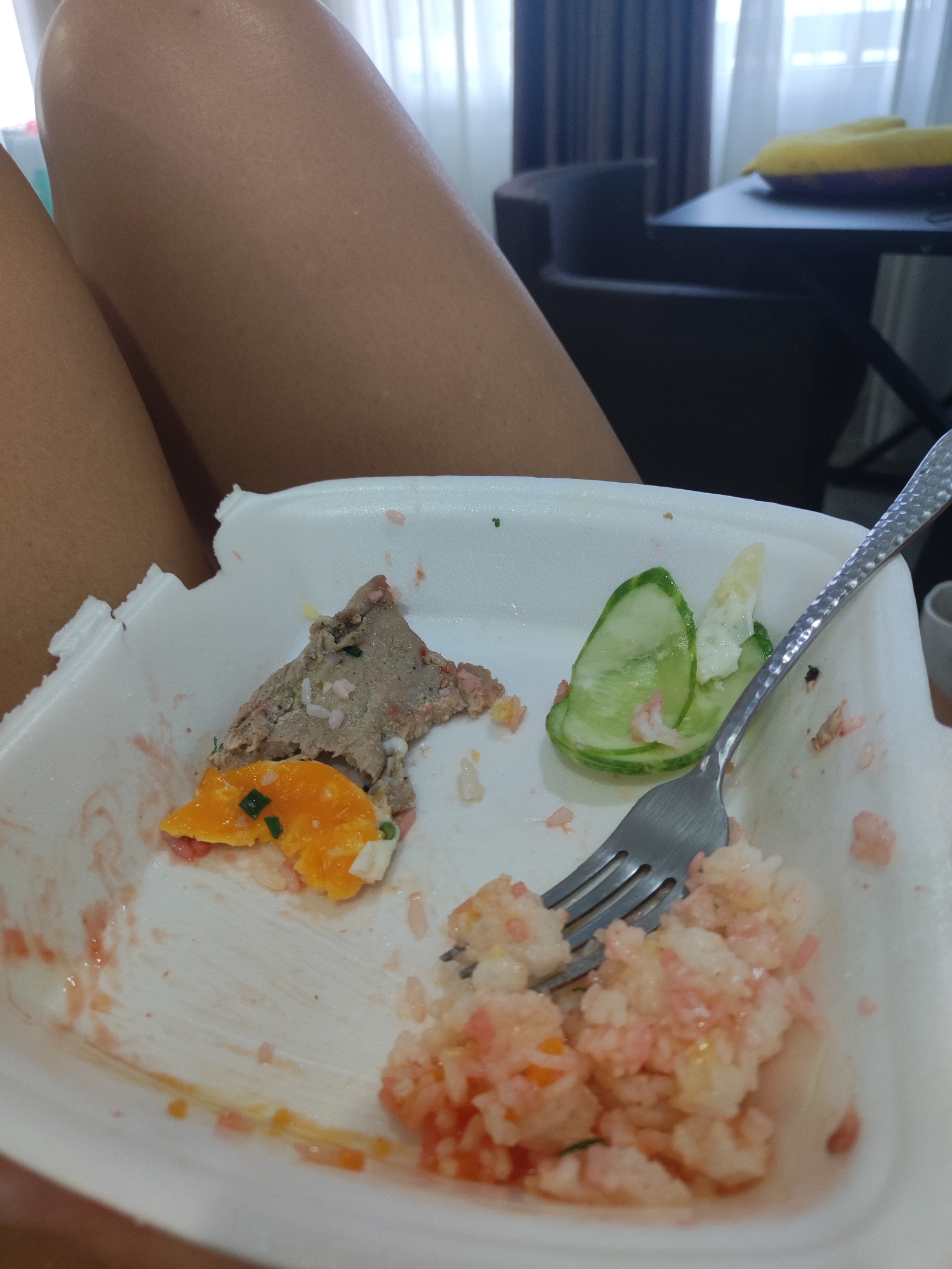 A close-up shot of a white foam container holding remnants of a meal. The container is partially filled with a fork, cucumber slices, some pinkish rice, and a piece of cooked egg yolk mixed with chopped green onions. A piece of meat or pate is also visible, with rice grains. The container itself is slightly crumpled. In the background, a person's legs are visible, and a blurred background suggests an indoor setting with curtains and furniture. The scene appears to be in a domestic or casual dining environment, likely in Nha Trang Ward, Vietnam, given the context.