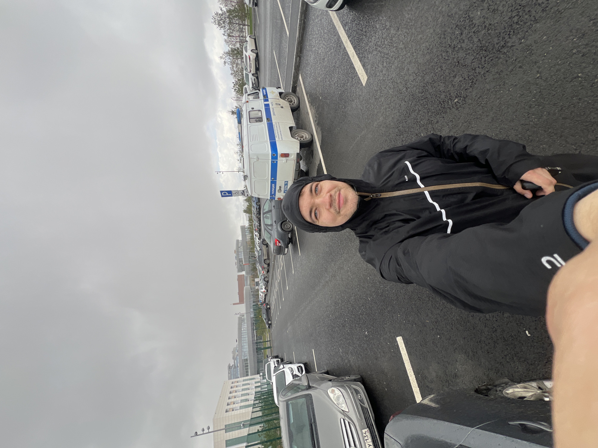 The image captures a man taking a selfie in a parking lot on a cloudy day. He is wearing a black hooded jacket and holding a small object in his hand. In the background, there is a police van parked in the lot, along with rows of other cars and a modern building. The license plate of a car in the foreground says "PY 134." The context of the image suggests it might be taken in Kazan, Russia, due to the markings on the police van and the license plate of the car.