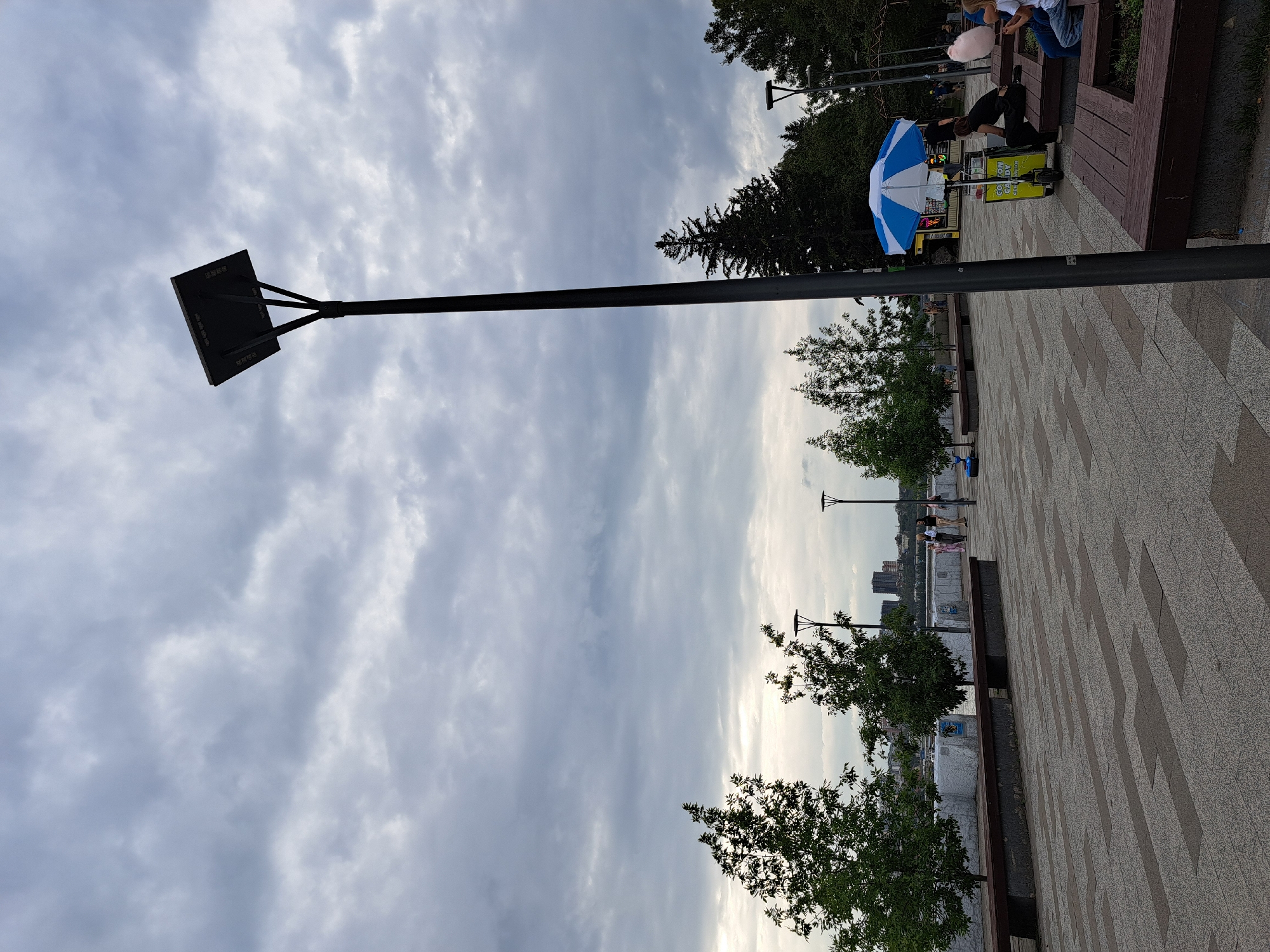 The image depicts a cloudy day on a waterfront promenade in Irkutsk, Russia. The scene is an outdoor public space with a paved walkway made of light and dark rectangular tiles. Benches line the promenade, some with people sitting on them. To the left, several young trees are planted along the edge, beyond which is a body of water and a cityscape with buildings. A tall, dark lamppost with a solar panel on top stands prominently in the foreground, angled towards the right. Further to the right, a cotton candy stand with a blue and white umbrella is visible, and a woman is seated on a bench nearby. In the background, evergreen trees form a dense line. The overall atmosphere is overcast and the light is diffused.
