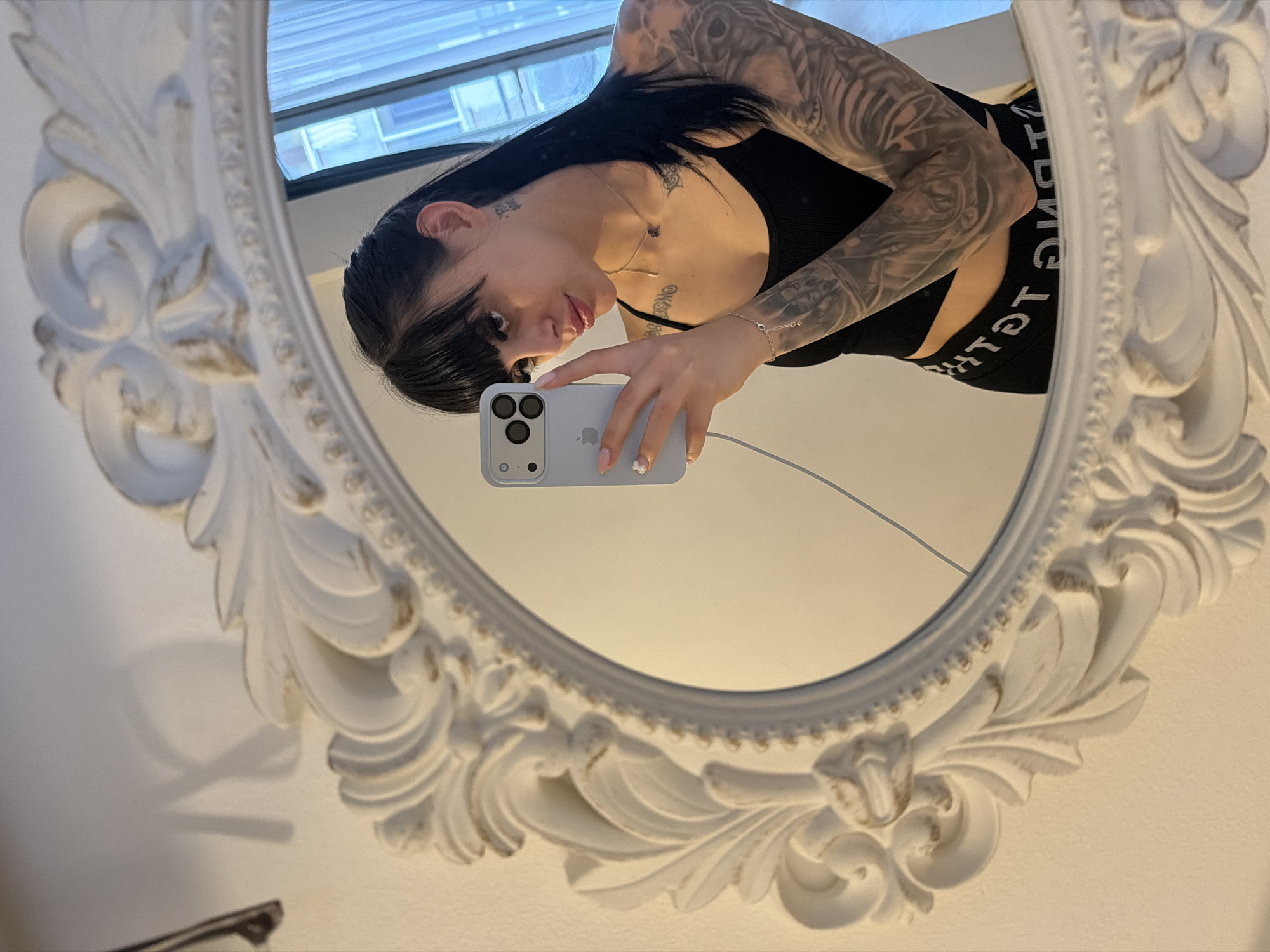 An indoor mirror selfie features a young adult female from the chest up, looking directly into the camera. She has long, dark hair with bangs and extensive black and grey tattoos covering both arms, depicting various designs including what appears to be a fierce animal. She is wearing a black sports bra or crop top and dark shorts, the waistband of which displays the mirrored text "STRENGTH". A delicate silver bracelet with small charms adorns her left wrist, and a simple necklace is around her neck.
She holds a light blue/grey iPhone with a triple-lens camera setup, connected to a charging cable. Her fingernails are painted light pink, with a white floral detail on one nail. The selfie is framed by an ornate, white, carved mirror with decorative scrollwork. Reflected in the mirror behind her is a window showing a glimpse of an urban environment or buildings, suggesting an indoor setting. The surrounding walls are light-colored. The lighting indicates it is likely daytime. There are no specific visual cues within the image itself to identify Plovdiv, Bulgaria.
