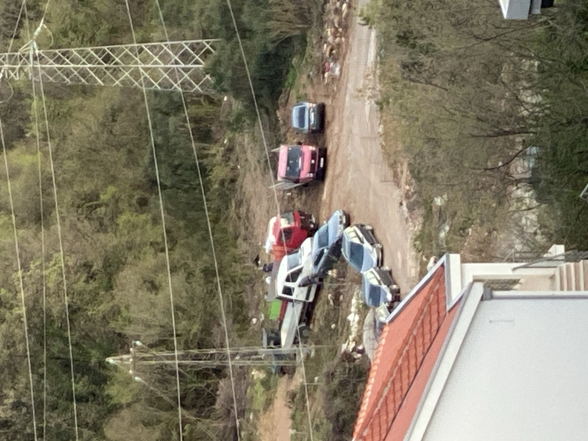 The image shows a clearing next to a dirt road near a wooded area, situated within the city of Budva, Montenegro. Several vehicles are parked in the clearing and along the dirt road, including two grey cars, a red car, a white truck, and a pink truck. Visible are portions of a building with a red-tiled roof and a white exterior. Overhead, a large power transmission line tower is prominently displayed. The environment consists of dense trees and vegetation surrounding the clearing and road.