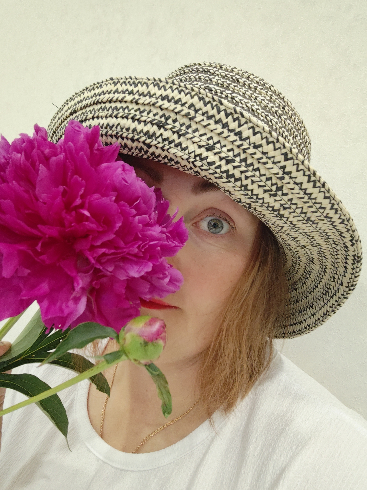 The image shows a woman in a straw hat holding a bright pink peony flower in front of her face. Her left eye is visible and is a light blue-gray color. She is wearing a white textured t-shirt and a delicate gold chain necklace. A portion of a peony bud is also visible. The background is a plain, light-colored wall. There is no indication of the city of Markovskii, Russia, in the image or its context.