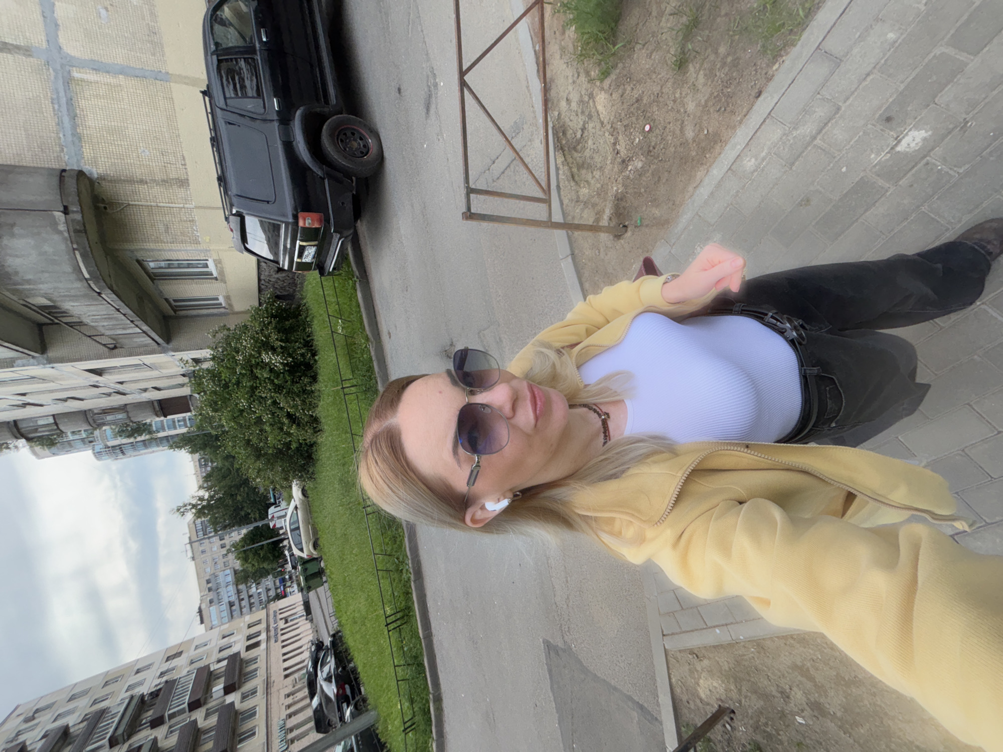 A blonde woman wearing sunglasses, a yellow jacket, a white top, and dark pants takes a selfie outdoors. She has white earbuds in her ears and a necklace around her neck. The background shows apartment buildings, parked cars, green grass, and a cloudy sky, consistent with an urban environment in Sinyavino, Russia. A black SUV is parked on the right side of the image. The ground is paved with grey concrete blocks.