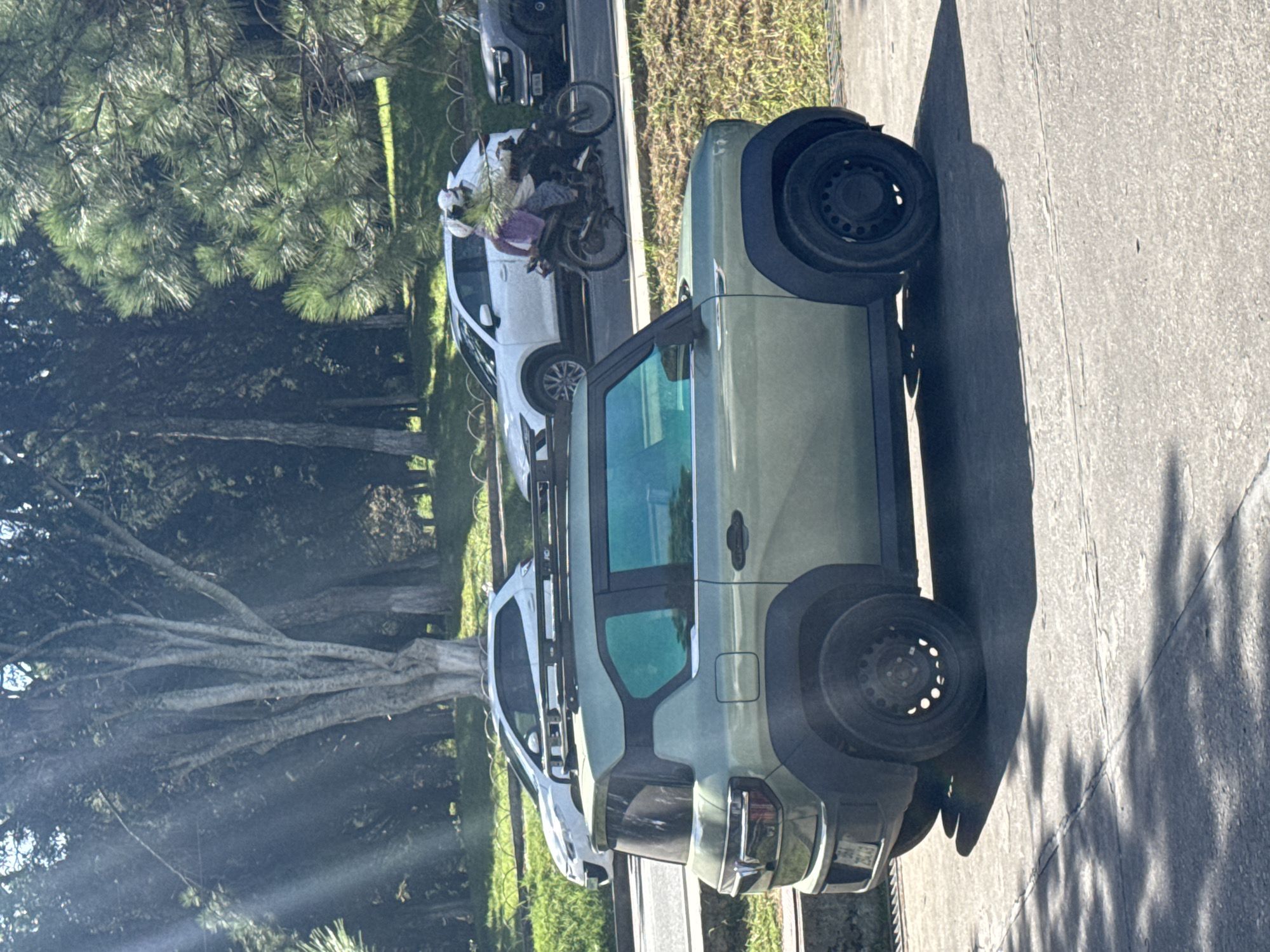 A greenish SUV with tinted windows and a roof rack is parked on a paved surface, casting a shadow. In the background, a white car is in motion, and a motorcycle with two occupants, one wearing a helmet and carrying a large bundle, is also moving. Beyond the vehicles, a grassy area with trees and a fence is visible. The image appears to be taken in Da Lat, Vietnam, given the lush greenery and distinct architecture. The sun's rays are filtering through the trees, creating lens flare effects.
