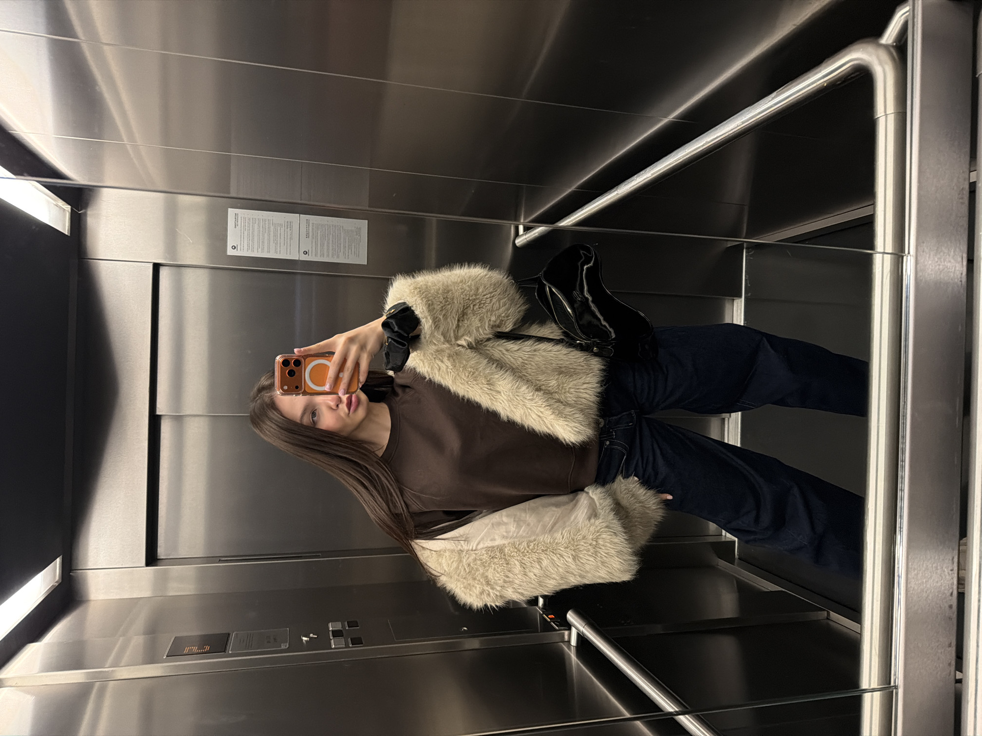 A young female is captured in a full-length mirror selfie inside a modern elevator. The elevator features brushed stainless steel walls, a dark ceiling with a long rectangular LED light fixture, and a dark, reflective floor. A stainless steel handrail is visible along the lower section of the side walls. A control panel with multiple buttons and a digital display is present on the left wall. Two white notices with Cyrillic text, confirming the location as Sofia, Bulgaria, are affixed to the right wall.
The individual has long, straight, dark brown hair and is wearing a dark brown long-sleeved top underneath a light beige faux fur jacket, worn open. She is dressed in dark blue, wide-leg denim jeans. A black scrunchie is on her left wrist, and a small, shiny black handbag with a distinctive asymmetric shape is carried on her left side. She holds an orange-cased smartphone with a circular MagSafe accessory in her right hand, extending her arm to take the photograph, and is looking directly at the camera.