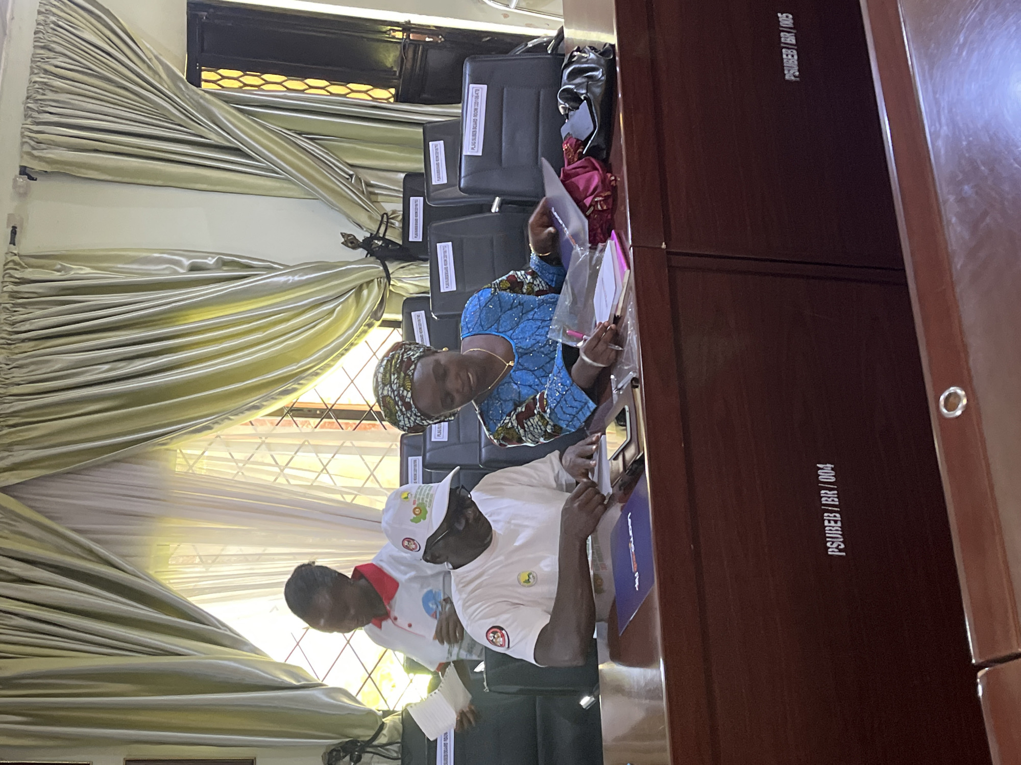 In the city of Jos, Nigeria, three individuals are seated at a dark wooden table in a room with light green and white curtains. In the foreground, a man wearing a white t-shirt and a white baseball cap with logos is writing on a document. To his right, a woman in a blue patterned dress and a headscarf is also looking at documents, with one hand holding a pen. Behind them, a person in a white polo shirt with a red collar stands and holds some papers. Several black chairs with white labels are visible in the background, as are two dark wooden cabinets in the foreground with labels reading "PSUBAB/BR/004" and "PSUBAB/BR/005". The overall scene suggests a meeting or administrative gathering.